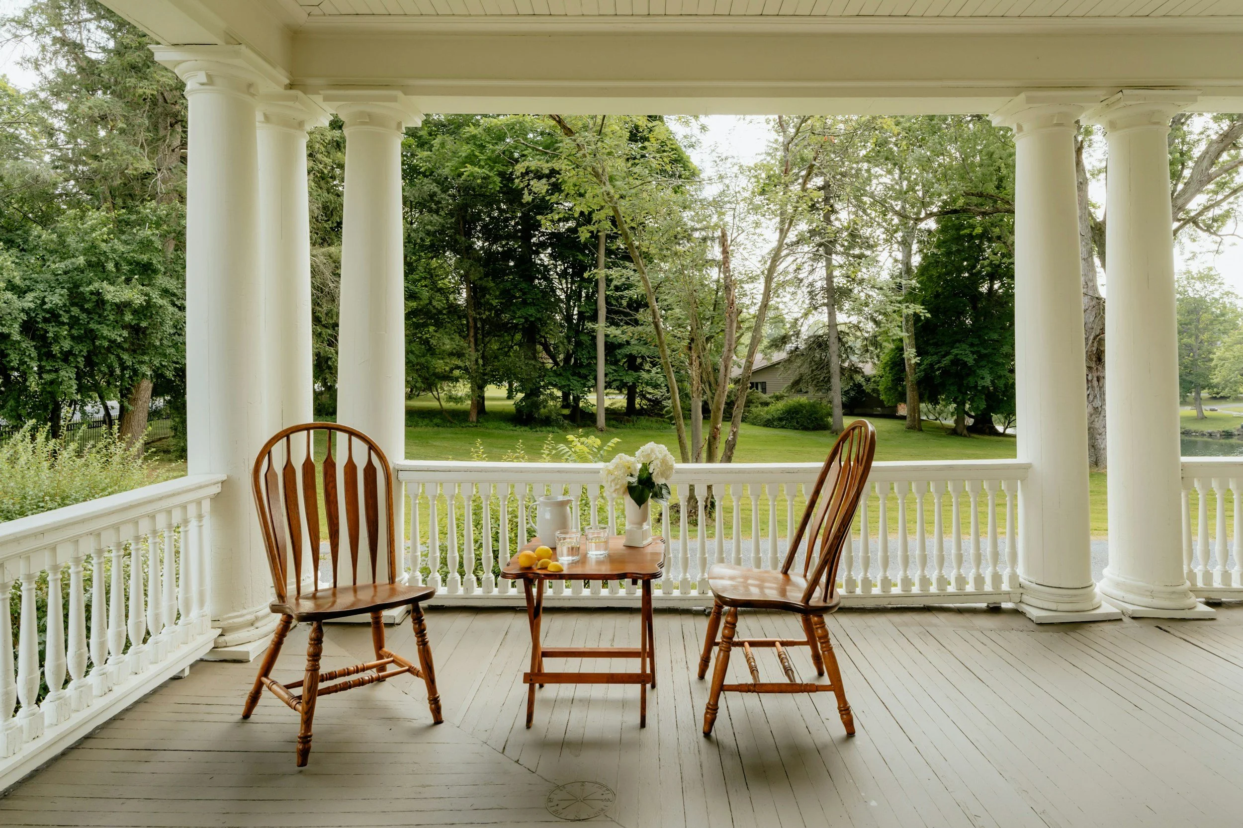A porch with white columns and railing overlooking a green lawn with trees. On the porch, there are two wooden chairs and a small wooden table with a vase of white flowers, a pitcher, glasses, and some fruits.