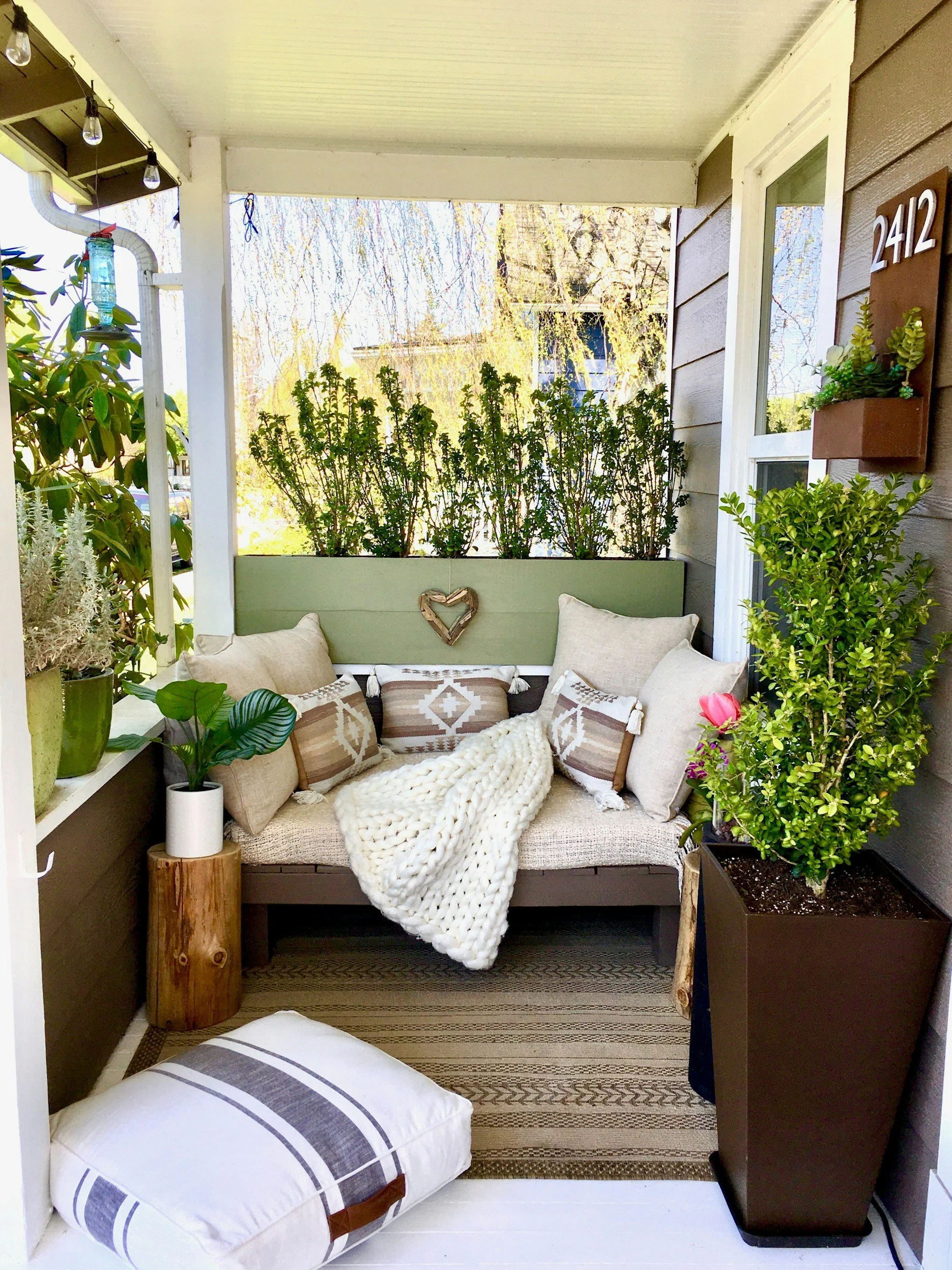 A cozy porch with a cushioned seating area surrounded by plants, decorative pillows, a knitted blanket, and a pillow on the floor. House numbers 2412 and a small succulent garden are visible.