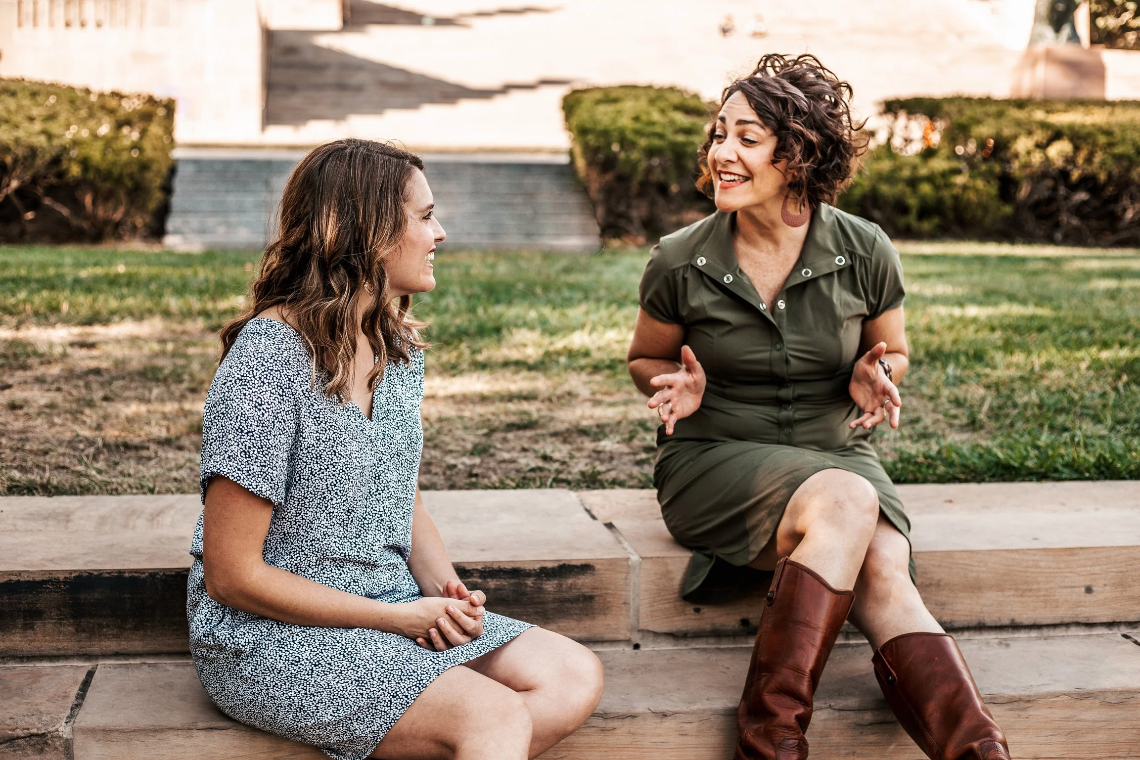 Two women sitting on a wooden step outdoors, engaged in conversation, with a grassy background and bushes.