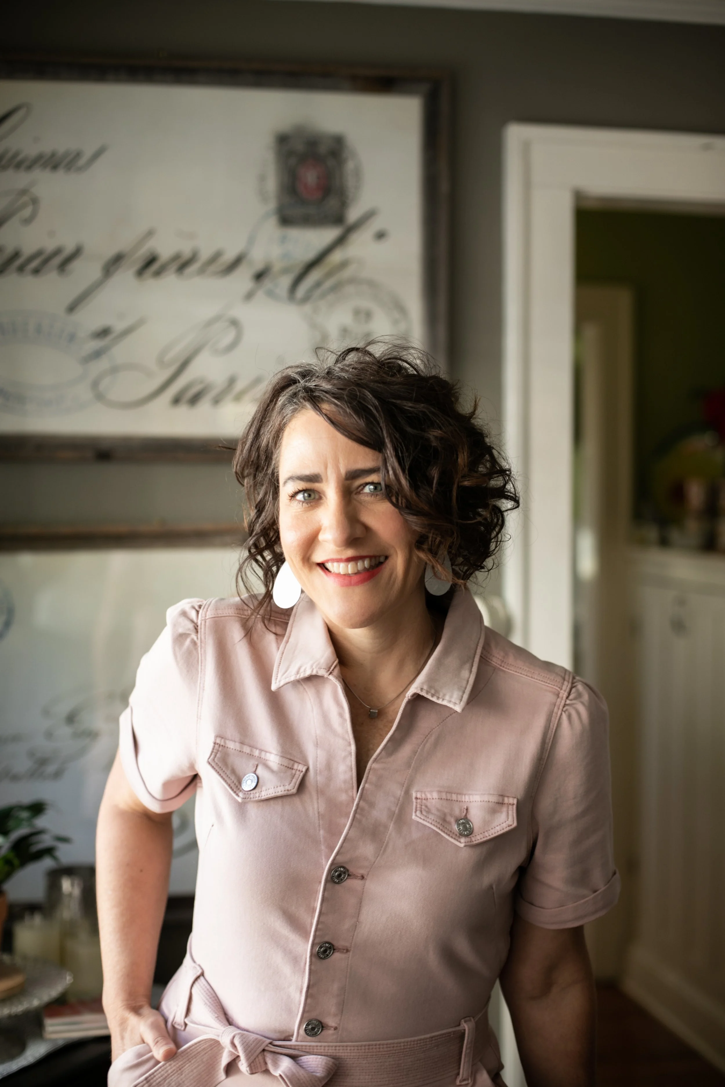A smiling woman with short, curly brown hair, wearing a light pink button-up dress with short sleeves, white teardrop earrings, and a delicate necklace, standing inside a home near a doorway with a blurred background.