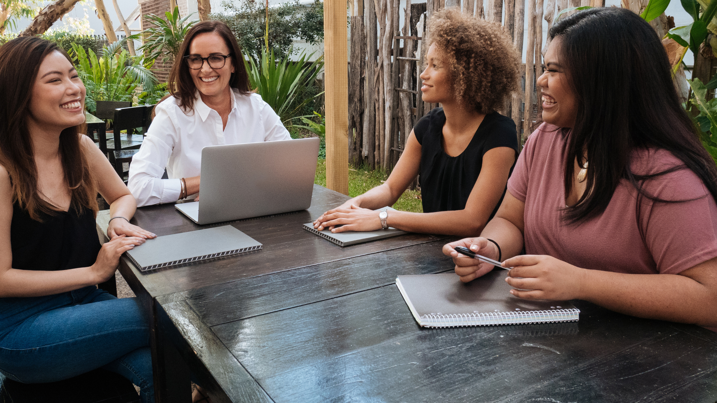 Four women sitting at a wooden outdoor table, smiling and laughing while having a discussion. One woman is using a laptop, and others have notebooks and one has a pen. Green plants and a wooden fence are in the background.