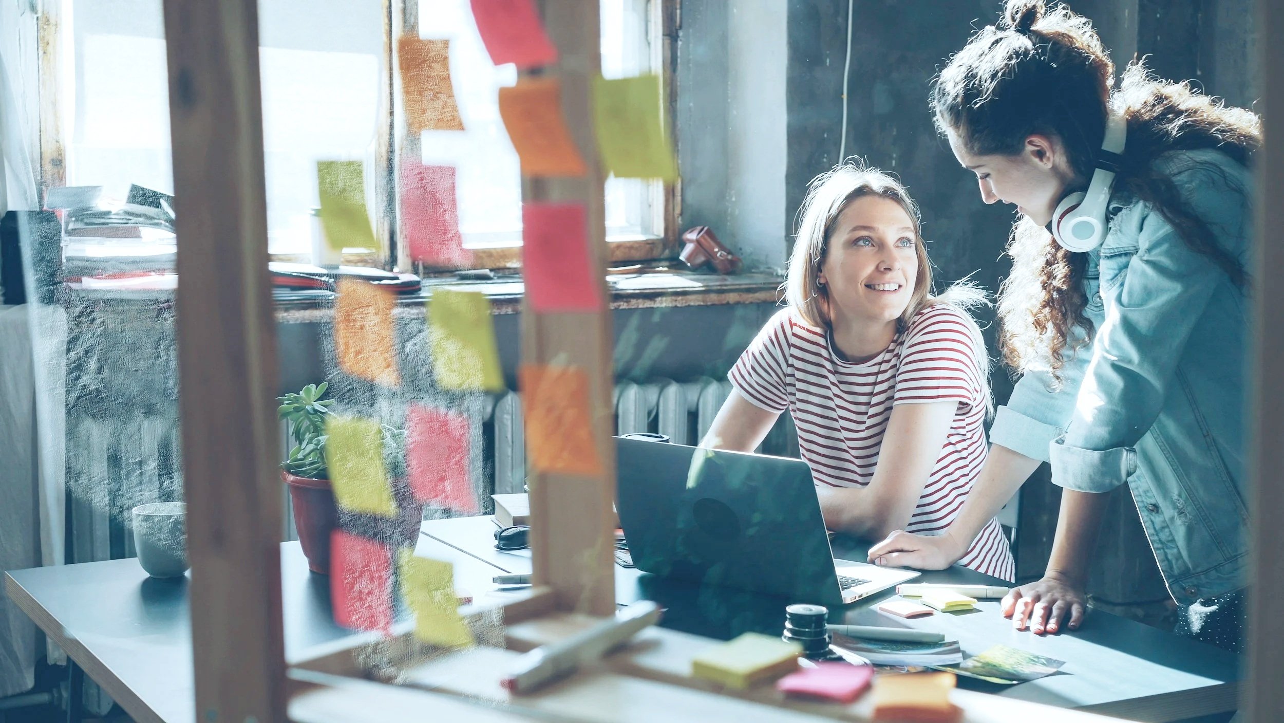 Two women working together in an office, one seated at a desk with a laptop, smiling, and the other standing with headphones around her neck, leaning over the desk, engaging in a discussion, with sticky notes on a glass wall in the foreground.