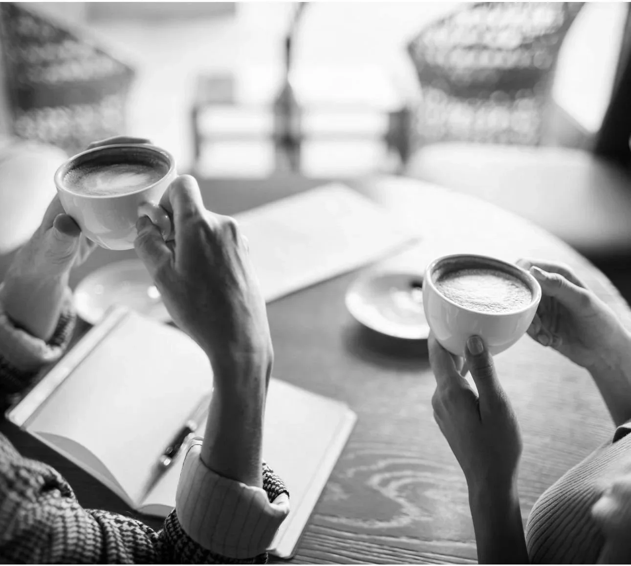 Two people holding cups of coffee or tea at a table, with an open notebook and pen nearby, in black and white.