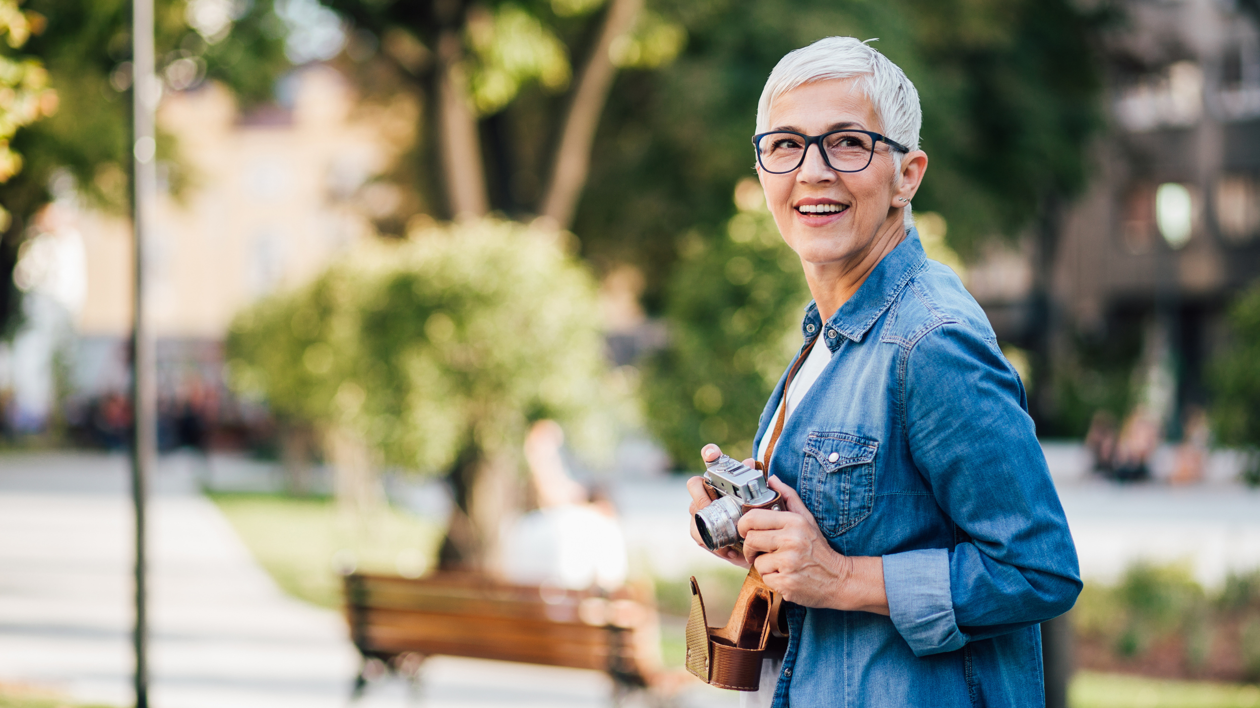 A smiling older woman with short gray hair, glasses, wearing a denim jacket, holding a vintage camera in an outdoor park setting.