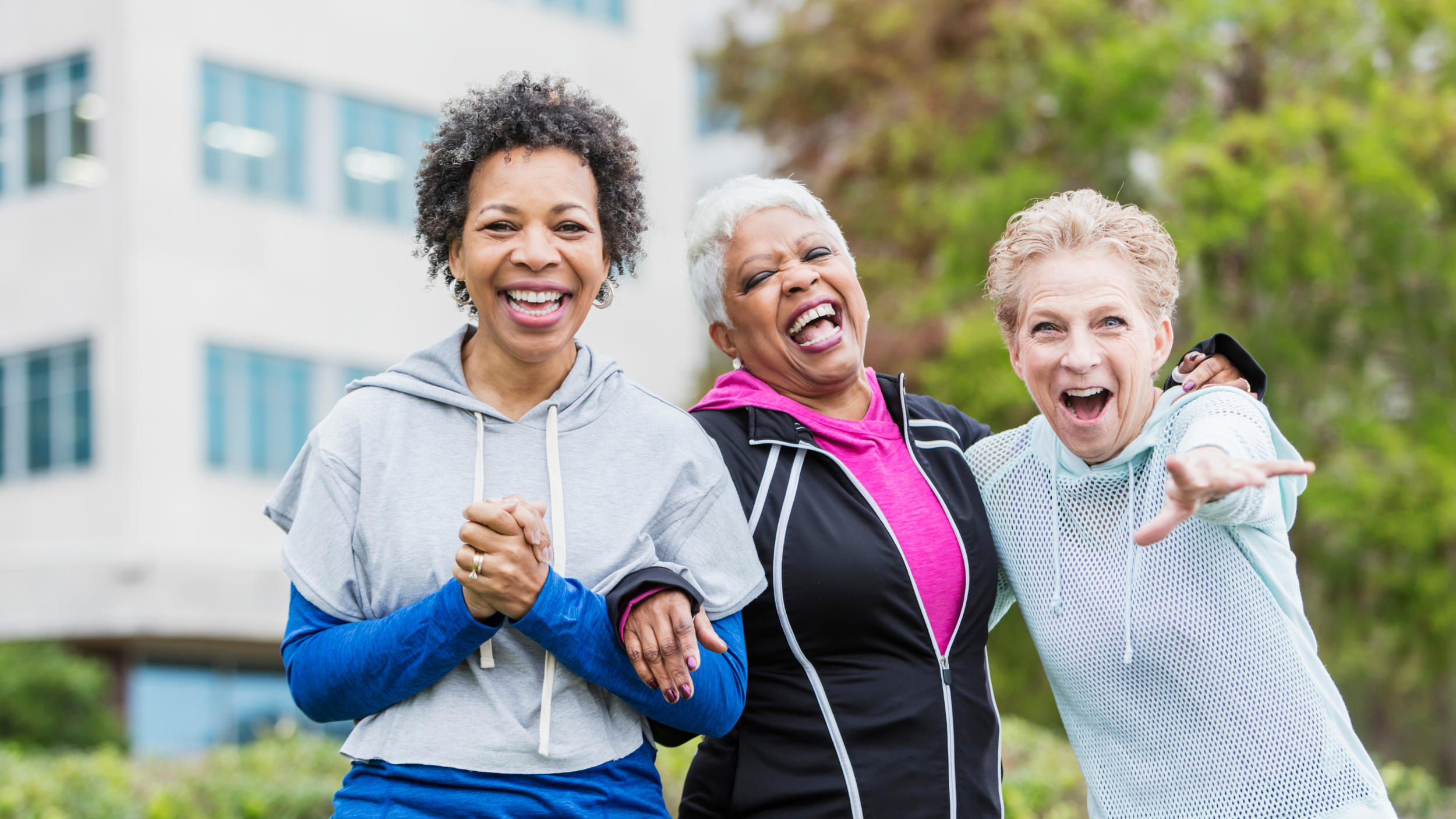 Three women smiling and cheering outdoors, with trees and a building in the background.