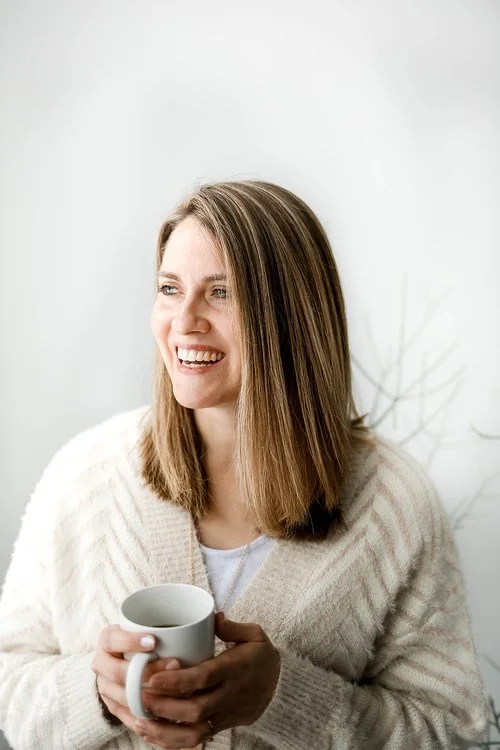 A woman with shoulder-length brown hair smiling and holding a white mug, standing against a plain white wall.