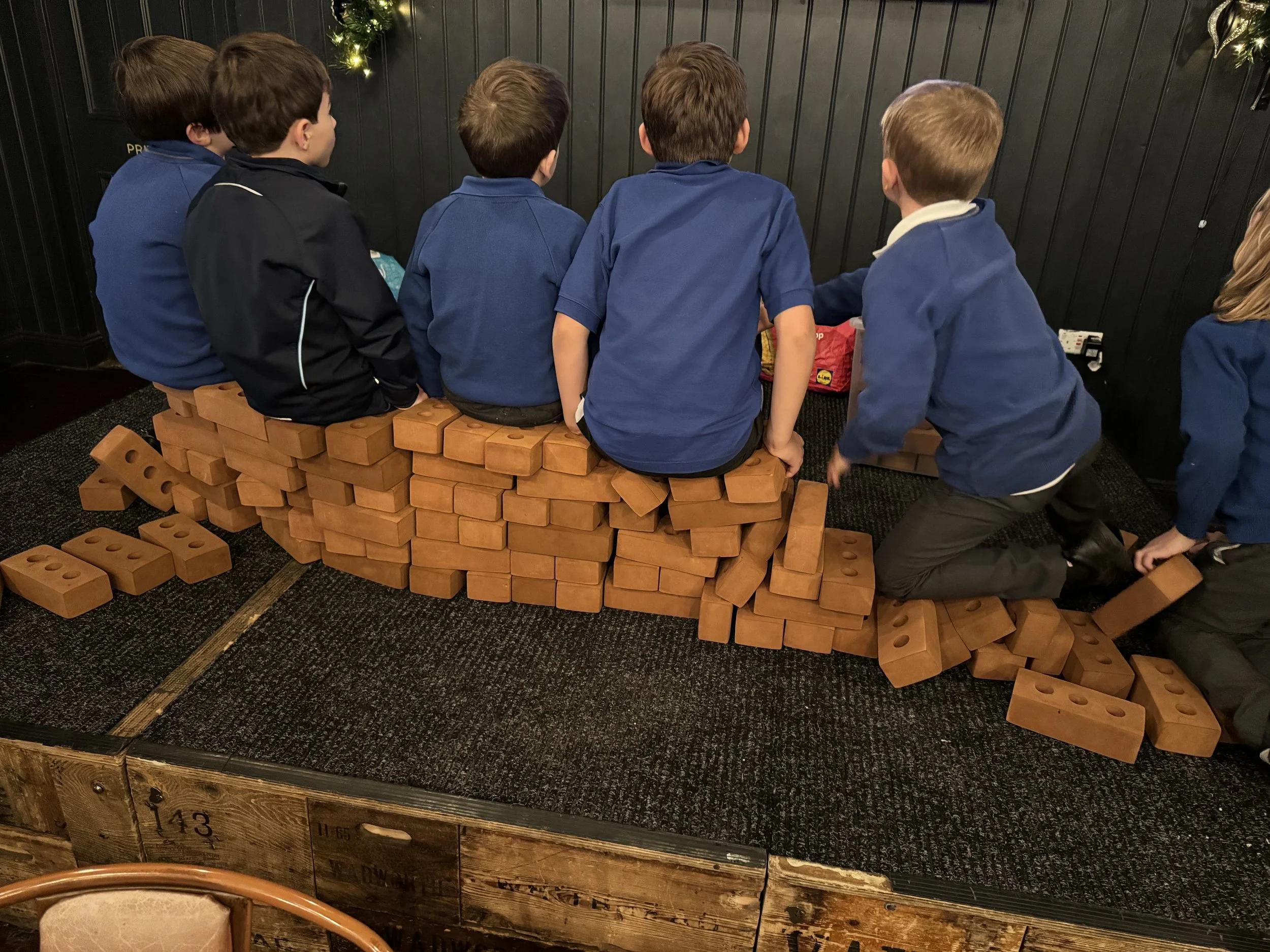 Children sitting on a stack of large toy bricks in a room with dark walls decorated for a holiday