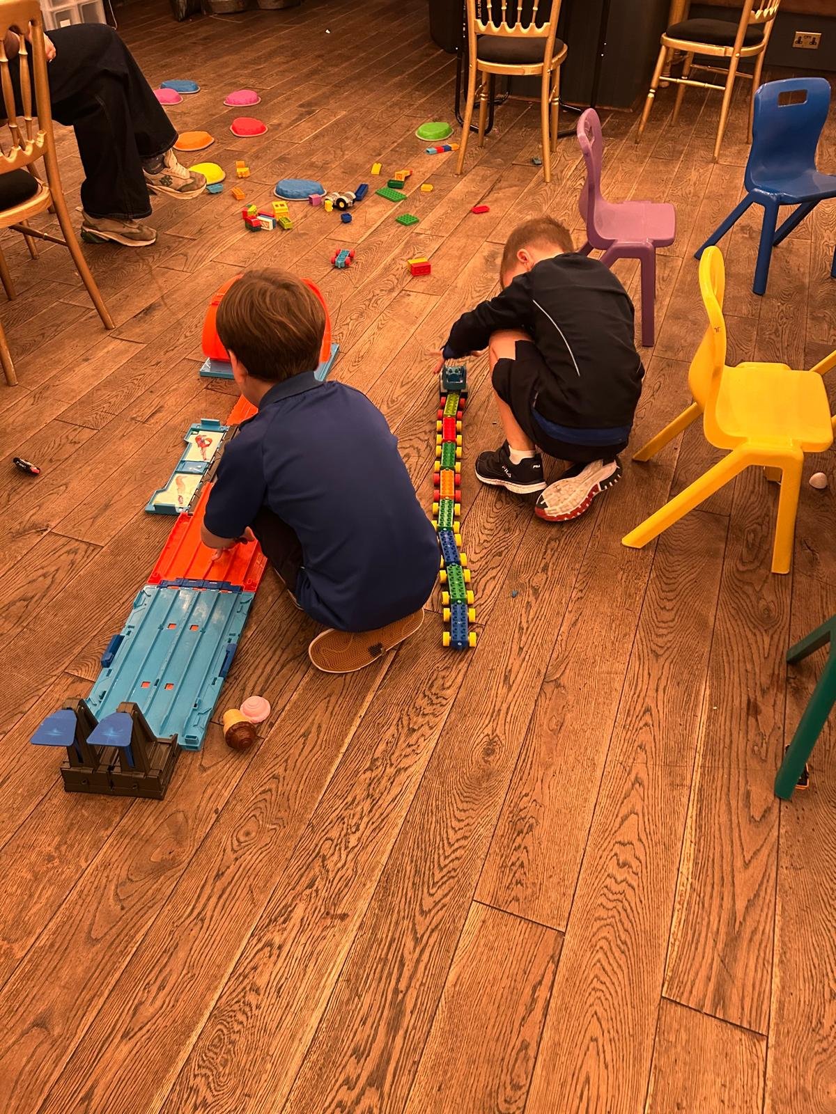 Two children sitting at a wooden table, surrounded by colorful yarn balls, with a brick wall in the background. 