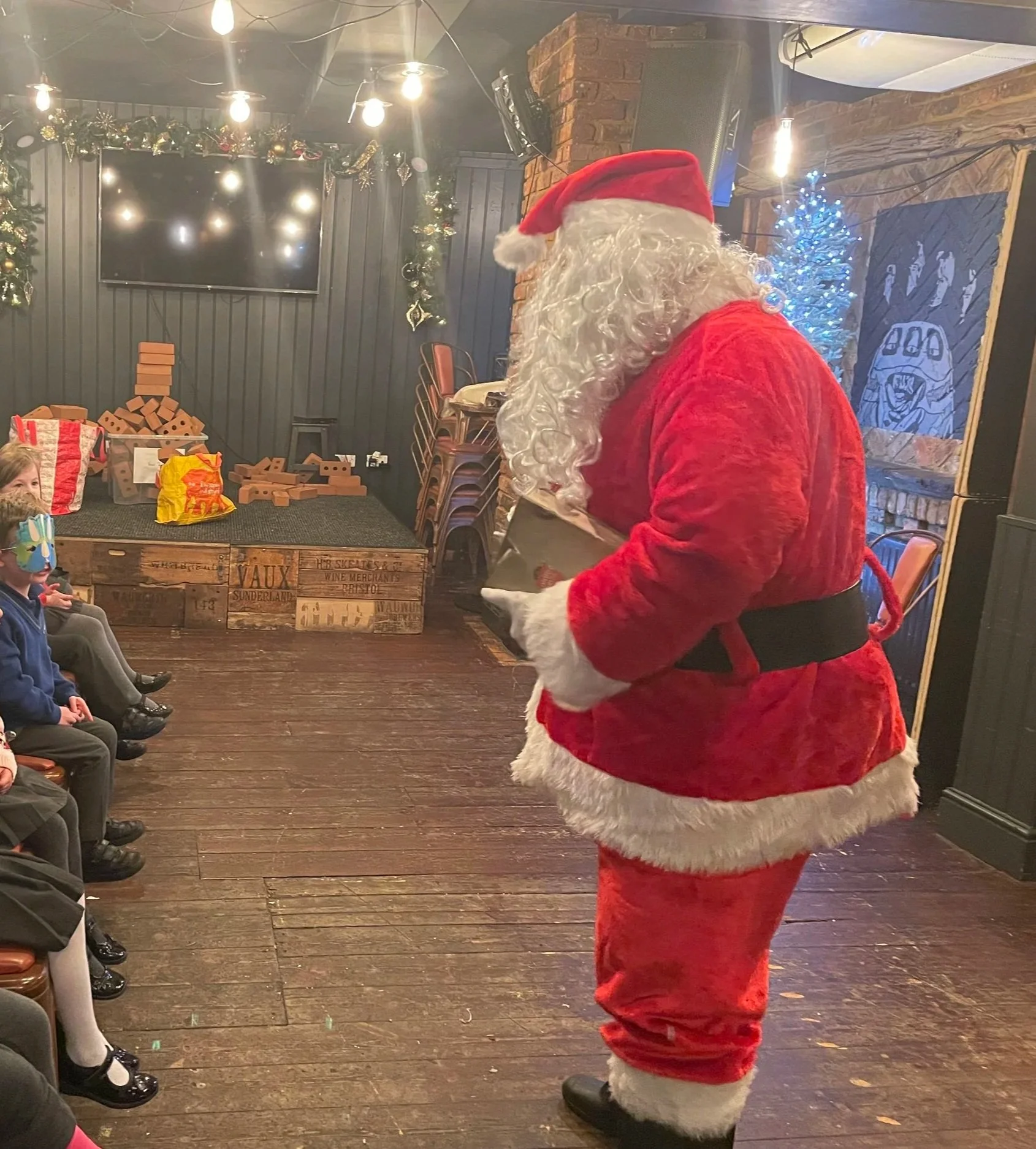 A group of children sitting in chairs, watching Santa Claus who is holding a book in a room decorated for Christmas with a small lit Christmas tree and wrapped gifts in the background.