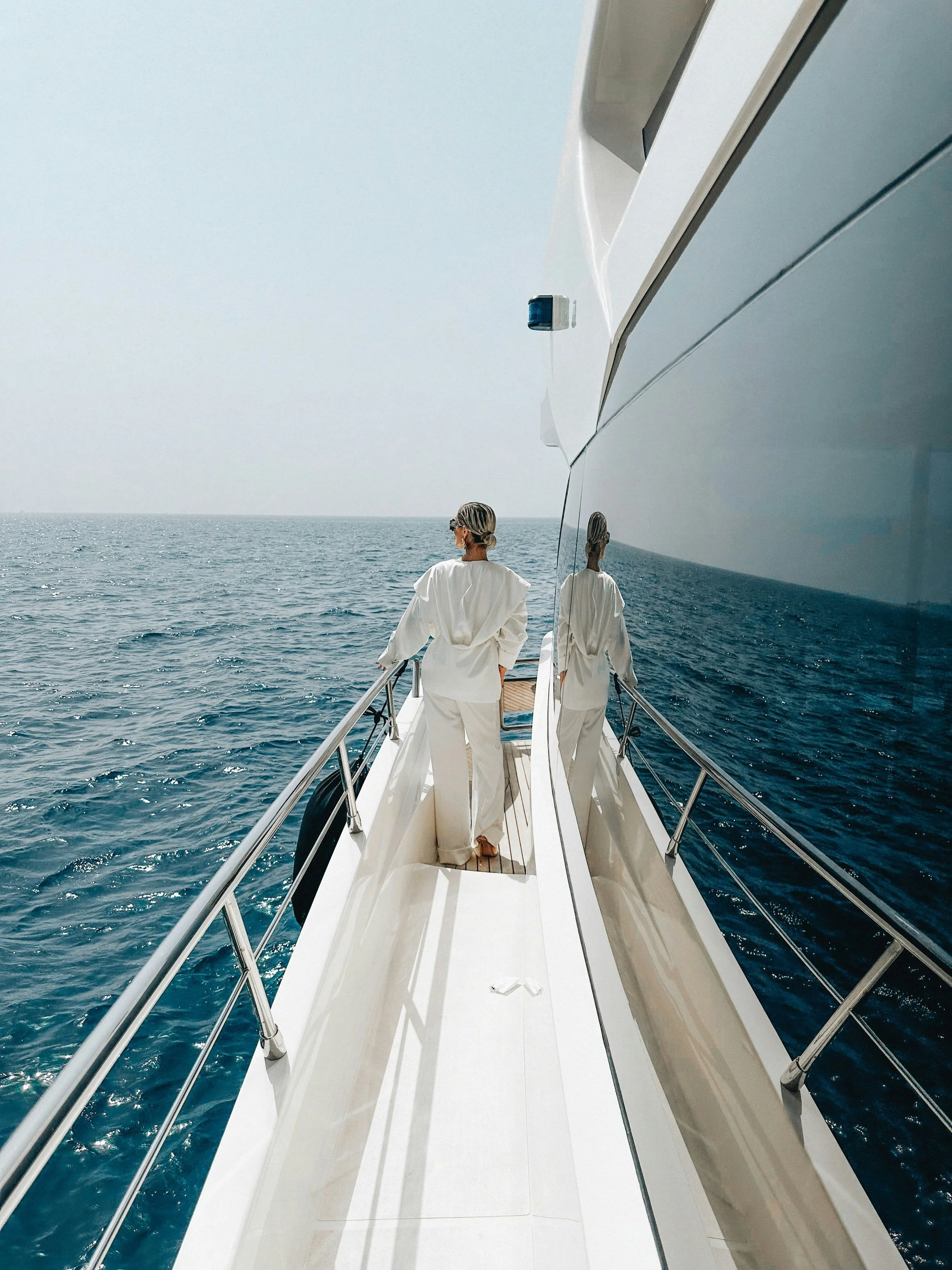 Person in white clothing walking on the deck of a yacht, with the ocean in the background and their reflection visible on the yacht's side.
