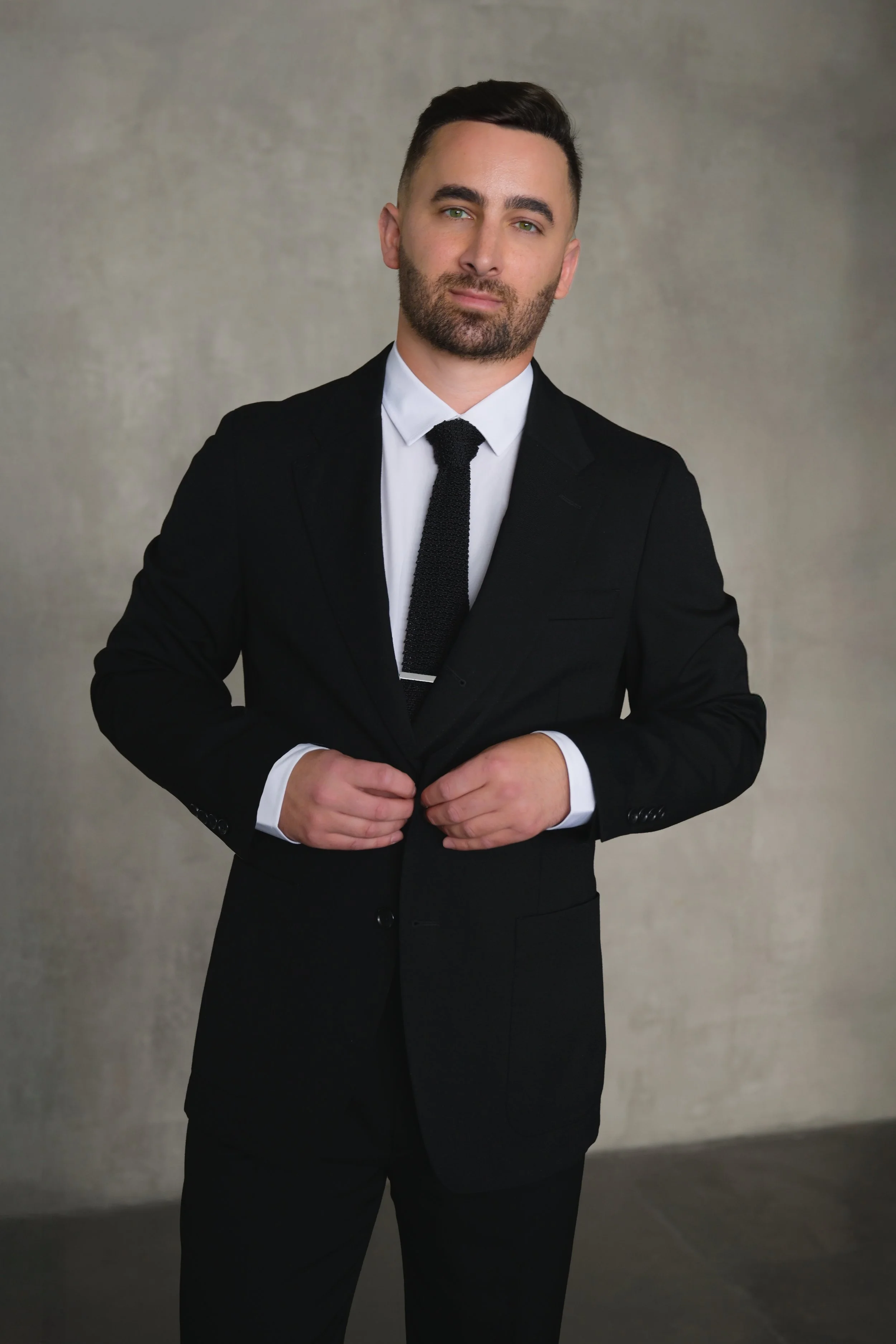 A young man in a black suit and white shirt adjusting his suit jacket, standing against a plain background.