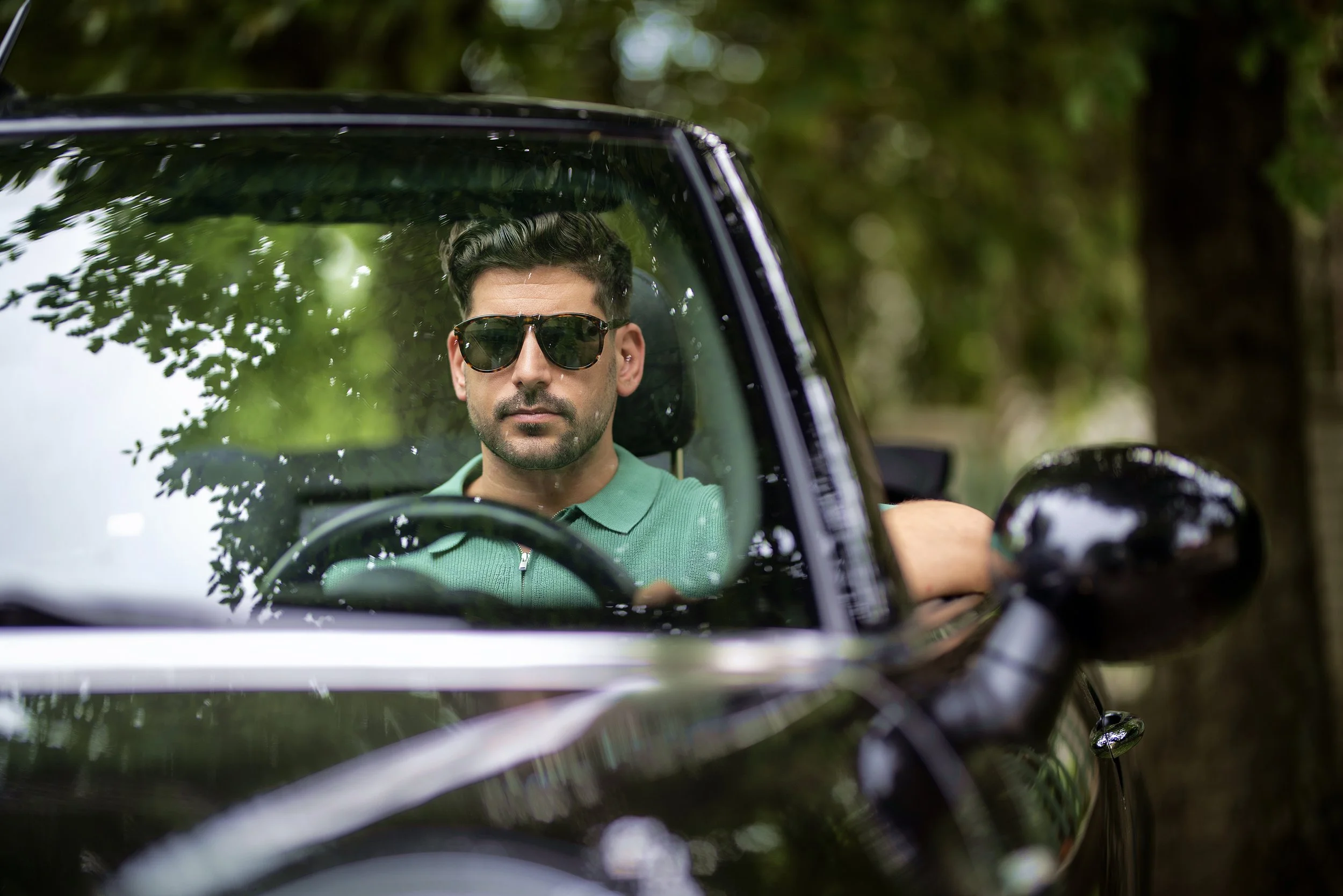 A man with dark hair, wearing sunglasses and a green zip-up shirt, sitting in a black car, viewed through the windshield, with trees and greenery in the background.