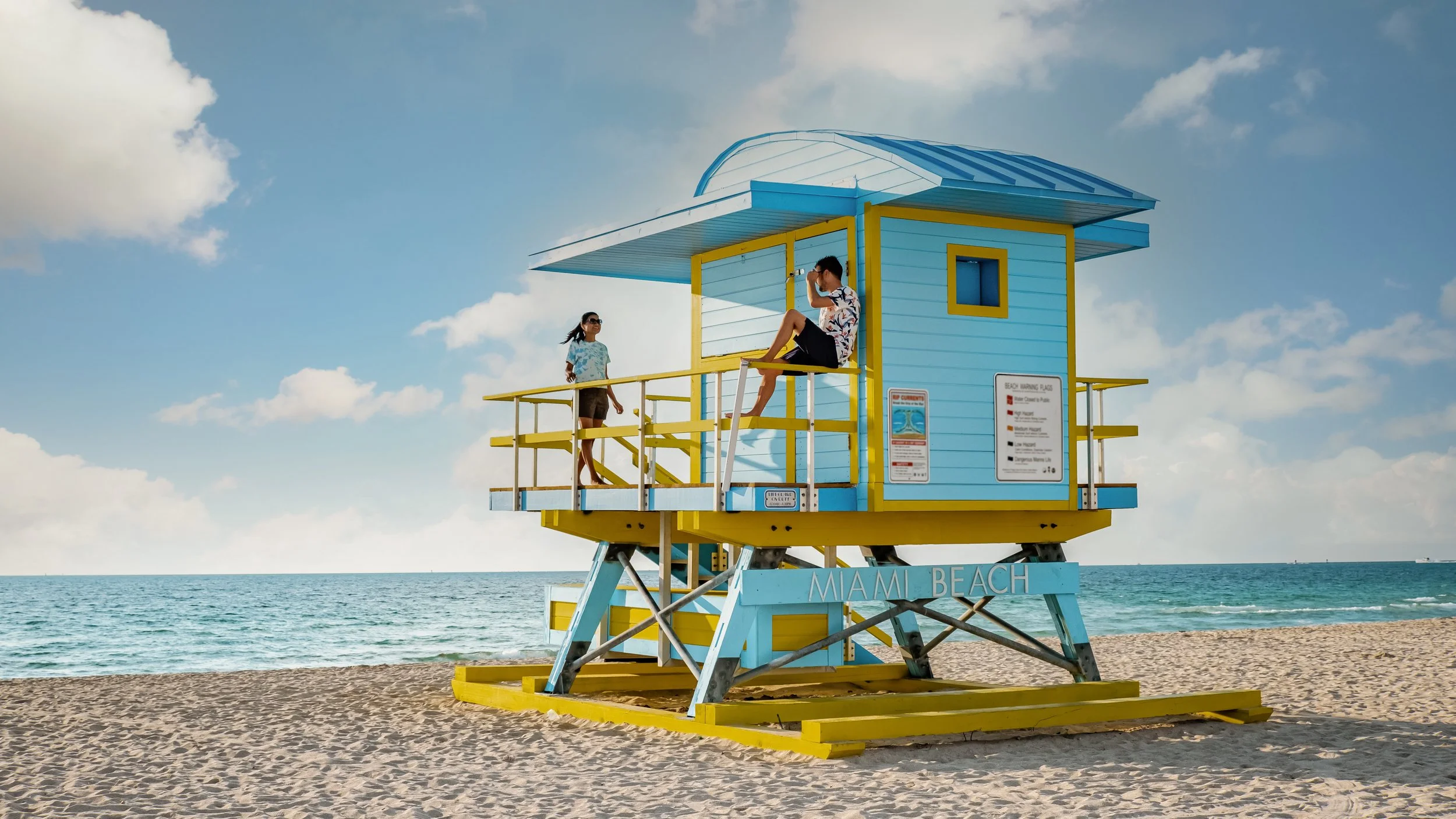 A colorful lifeguard tower on Miami Beach with three people on it, overlooking the ocean and a blue sky with clouds.