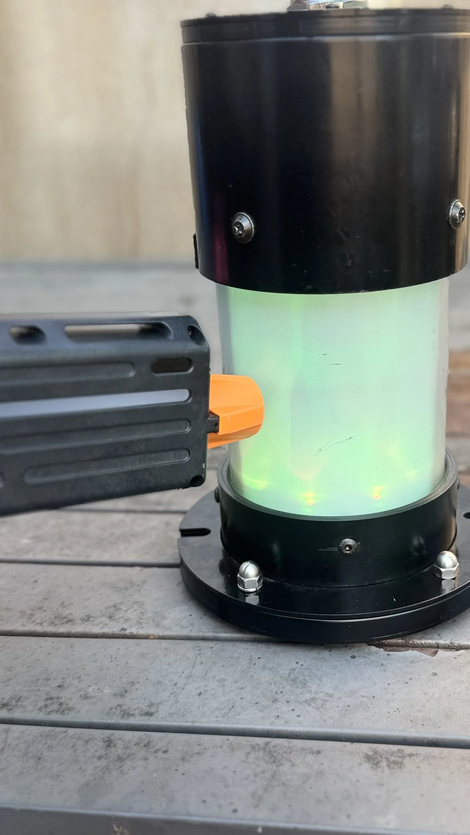 A black and clear cylindrical device emitting green light with electrical components attached, placed on a wooden surface.