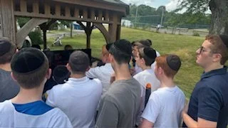 Group of boys and young men standing outdoors, facing a wooden structure, watching something or listening attentively.