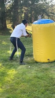 Person participating in a paintball game, taking cover behind a large yellow inflatable obstacle outdoors.