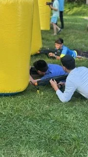Three boys lying on the grass and aiming toy guns at a yellow inflatable structure during a daytime outdoor activity.