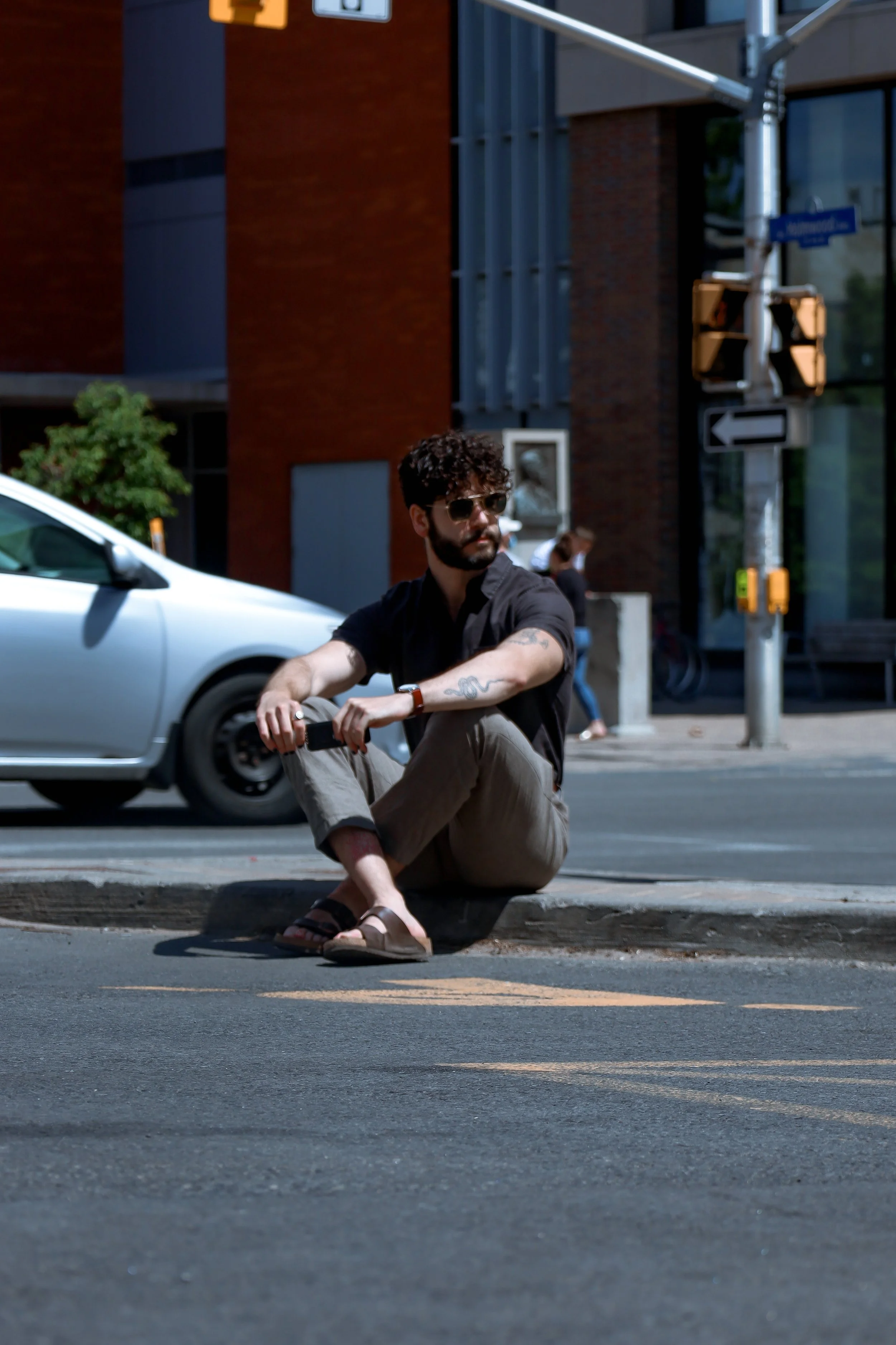 A young man with curly hair, beard, sunglasses, and tattoos, sitting on the curb at a city intersection during daytime, wearing a black shirt, beige pants, and sandals.