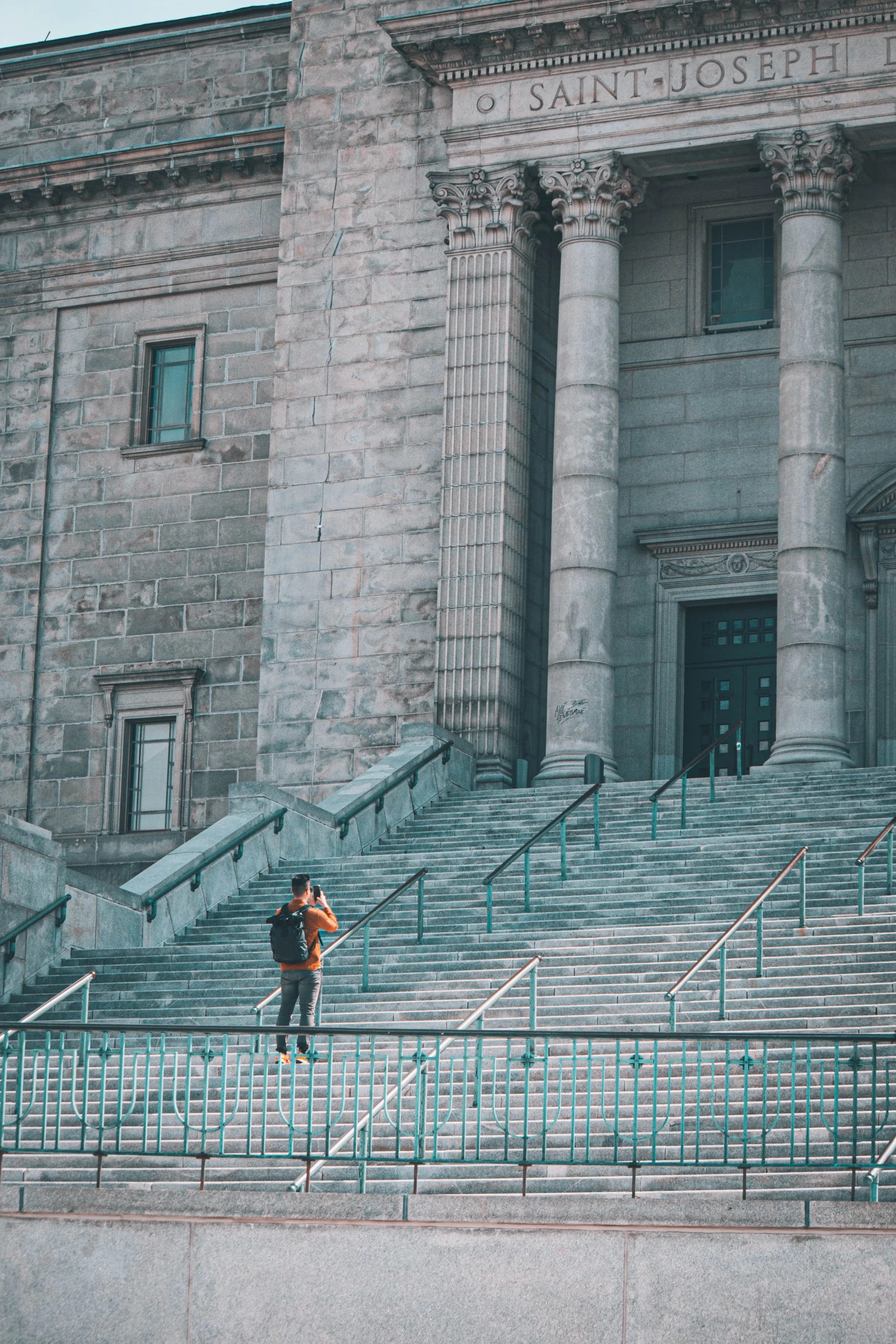 A person standing on the stairs outside a large stone building with the sign 'Saint Joseph' and taking a photo.
