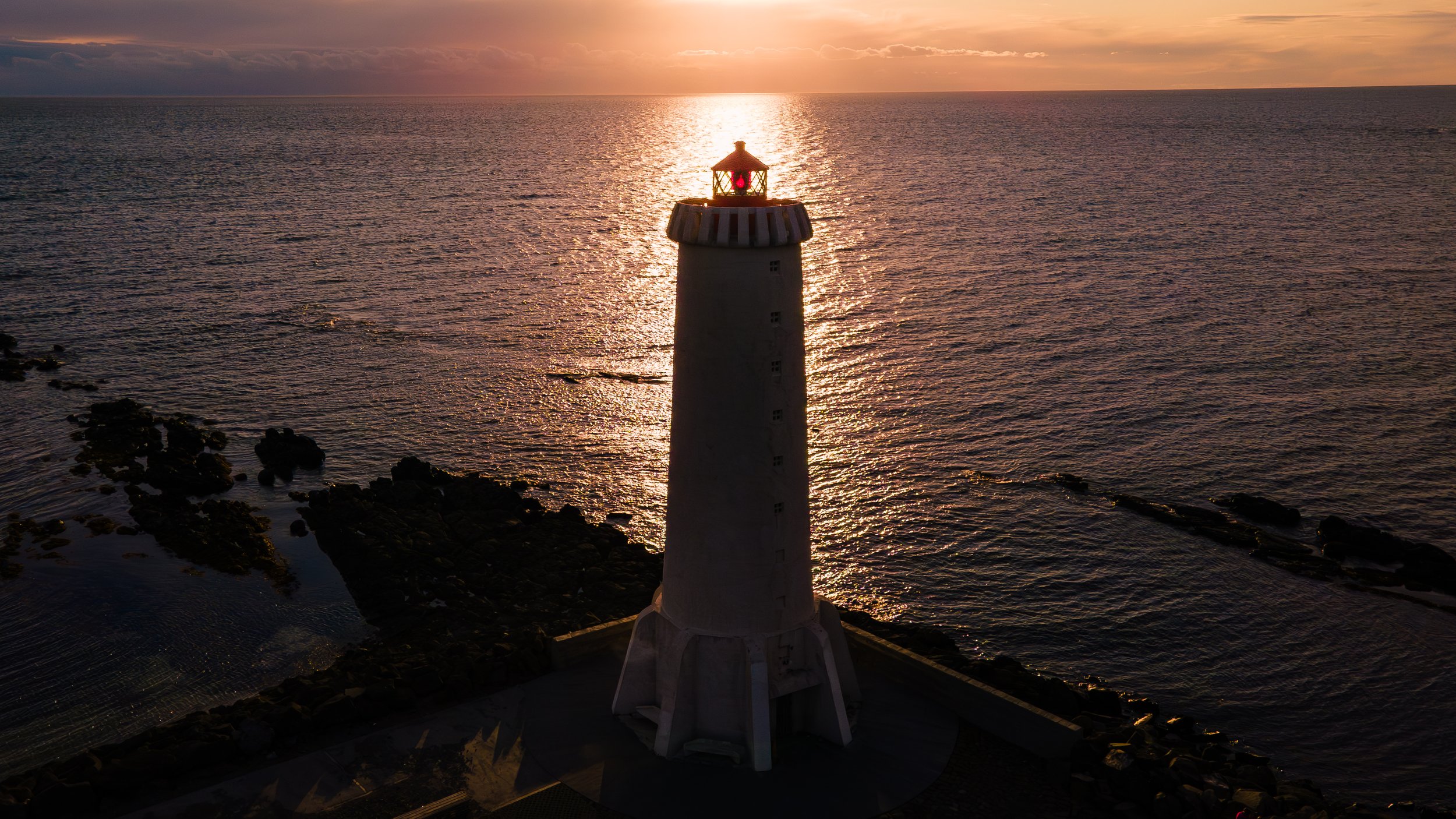 Lighthouse on a rocky coast during sunset, with the sun reflecting off the water.