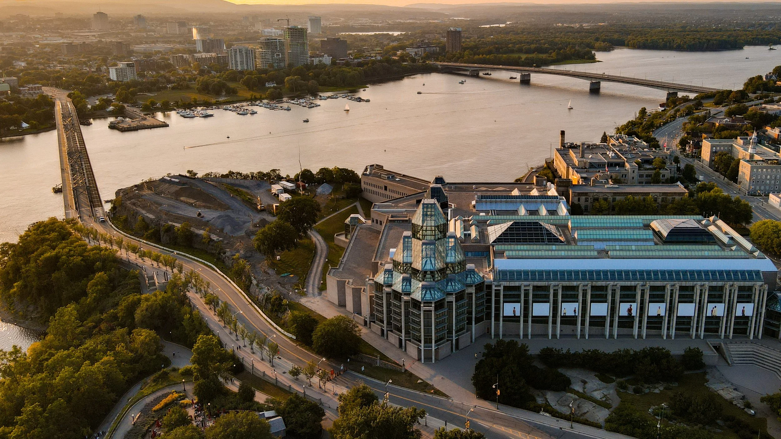 Aerial view of a cityscape featuring a large building with a glass roof and towers, a river with boats, a bridge, and a surrounding urban area at sunset.