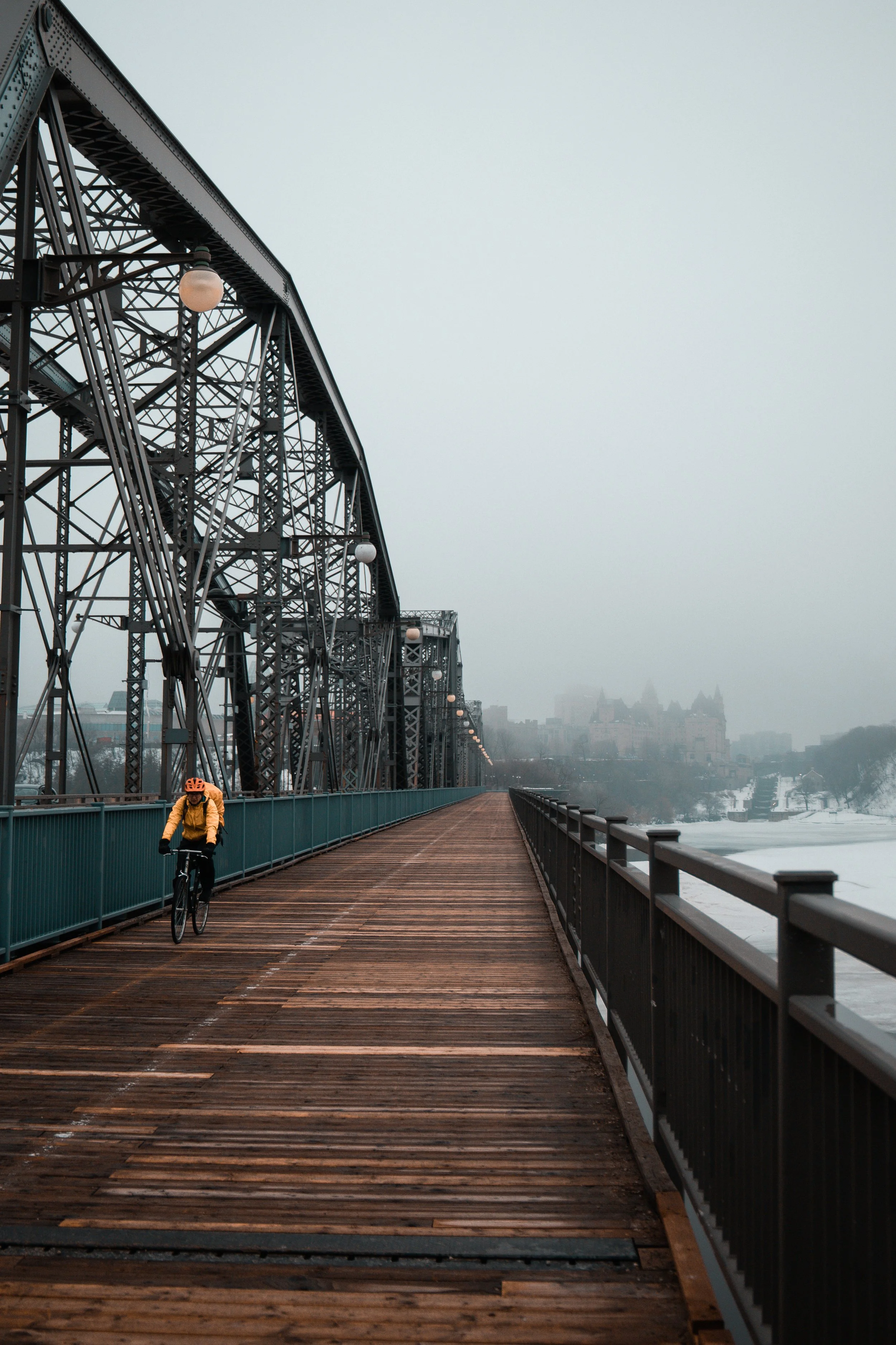 A cyclist in an orange helmet and yellow jacket riding on a wooden bridge over a frozen river, with a city skyline and a castle-like building in the background on a cloudy day.