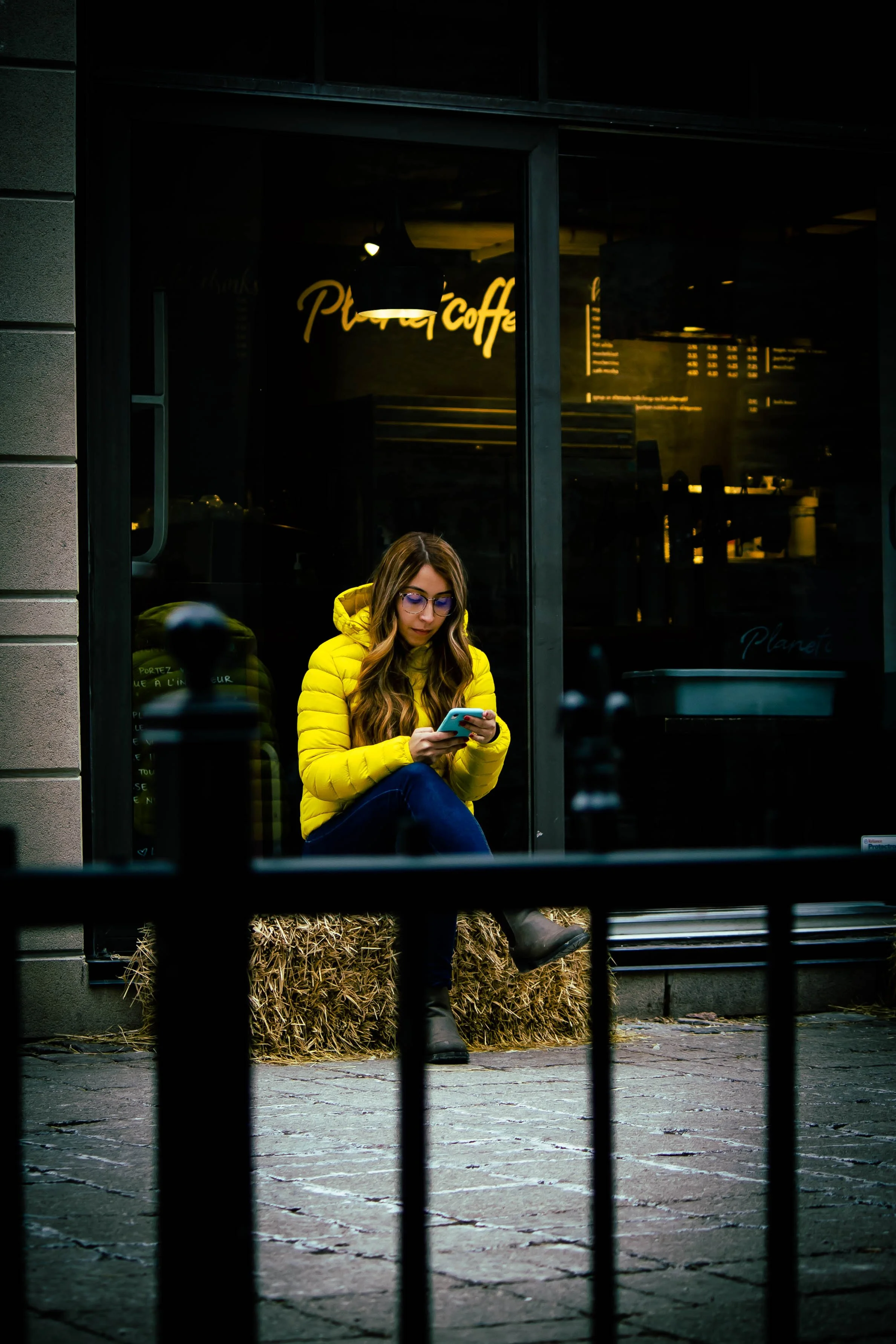 A woman in a yellow jacket sitting on a hay bale outside a cafe, looking at her phone. The cafe window behind her has a neon sign in cursive that says 'Planet Coffee', and the scene is framed by a black metal fence in the foreground.