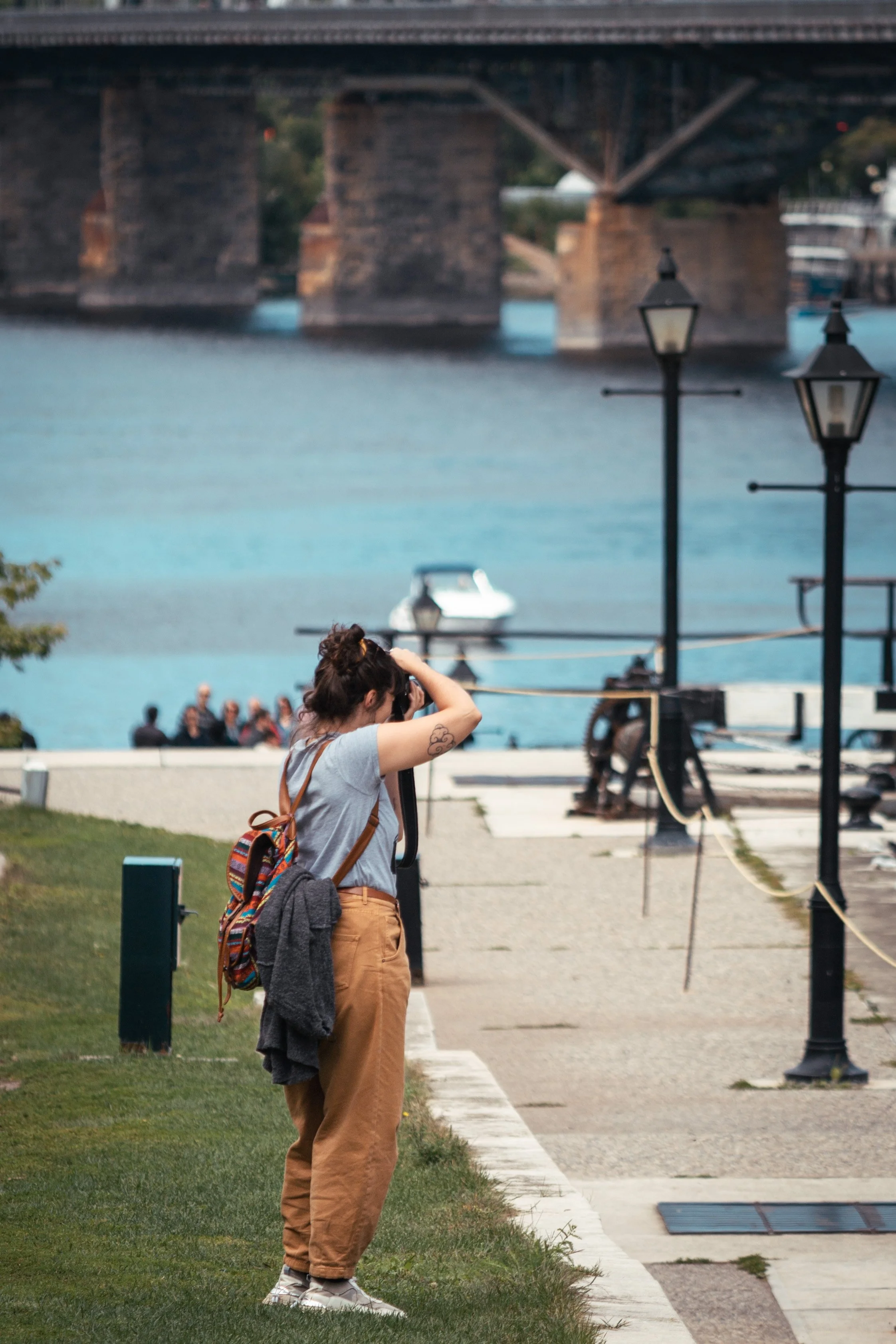 A woman with a bun hairstyle, carrying a colorful striped backpack, standing on a grassy area near the water, taking a photo with a camera. There are lamp posts, a dock, and a boat in the background.