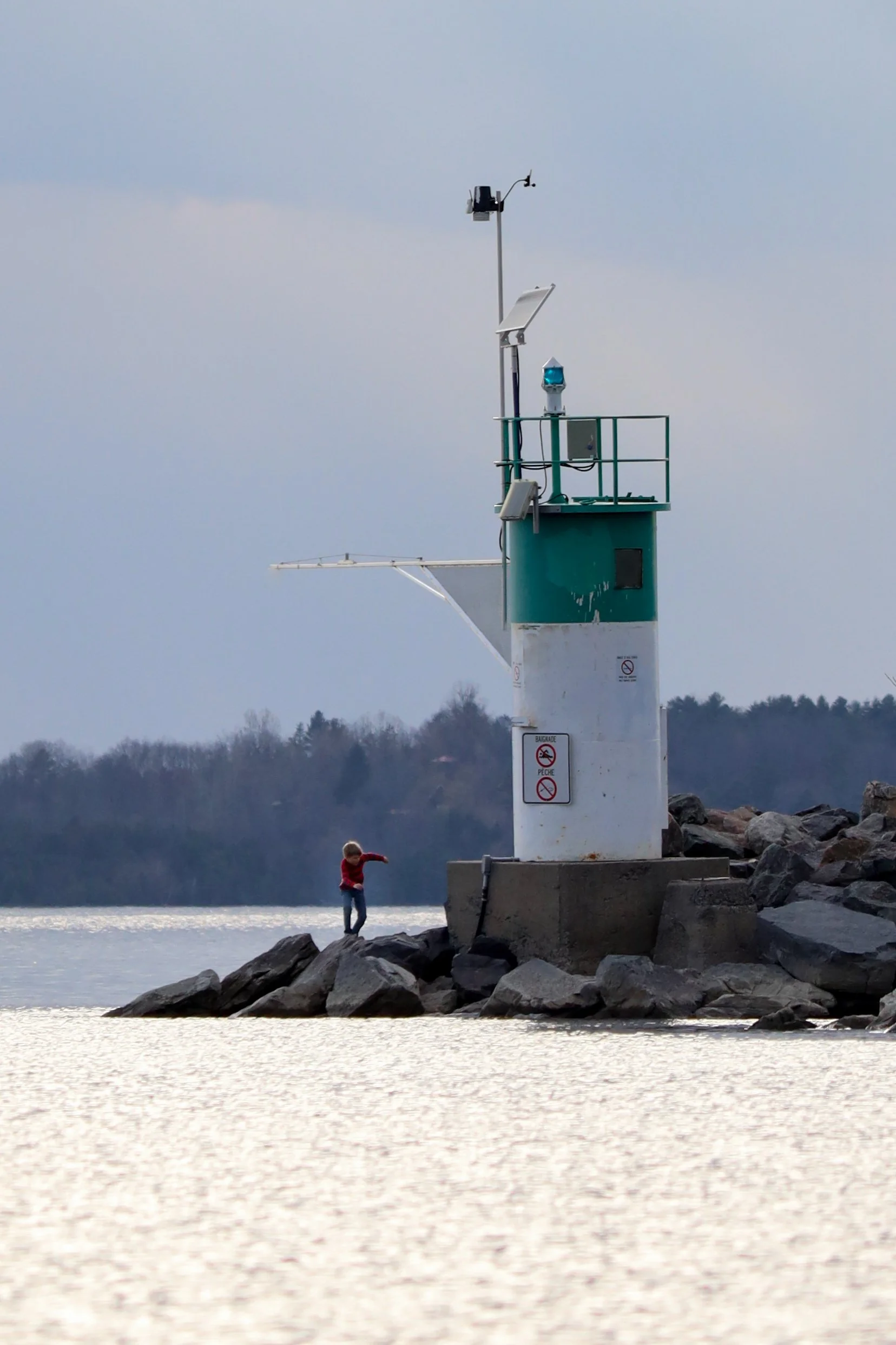A small child in a red jacket standing on rocks near a lighthouse on a shoreline, with water and a distant tree line in the background.