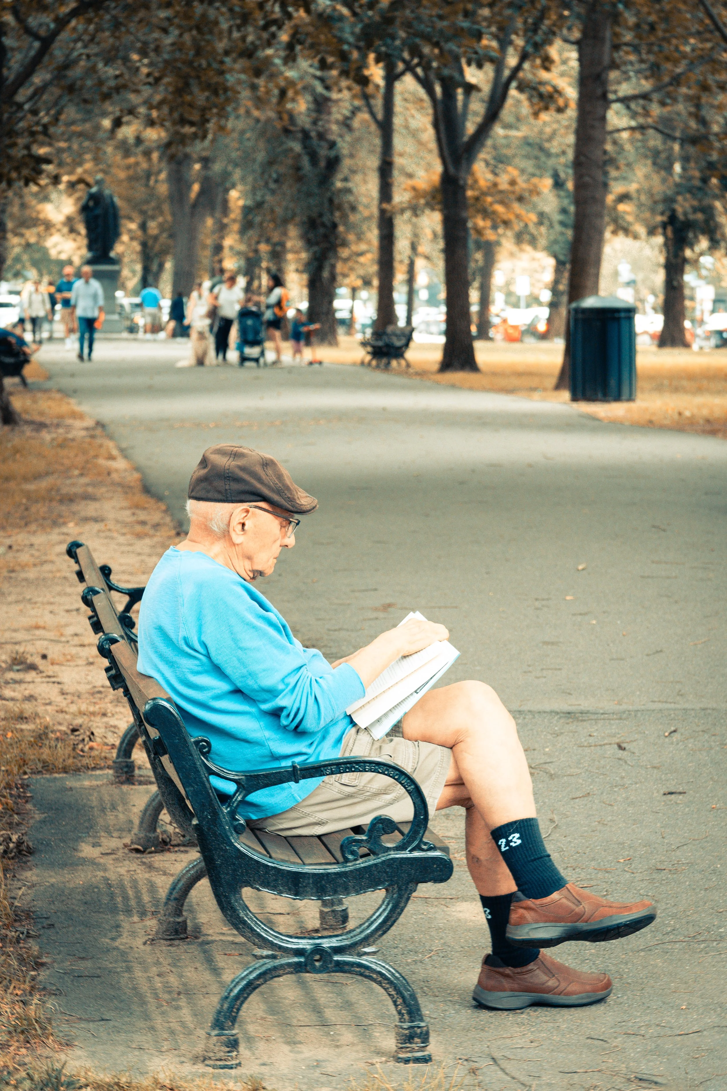 An elderly man sitting on a park bench reading a newspaper, surrounded by trees with autumn foliage and other park visitors in the background.