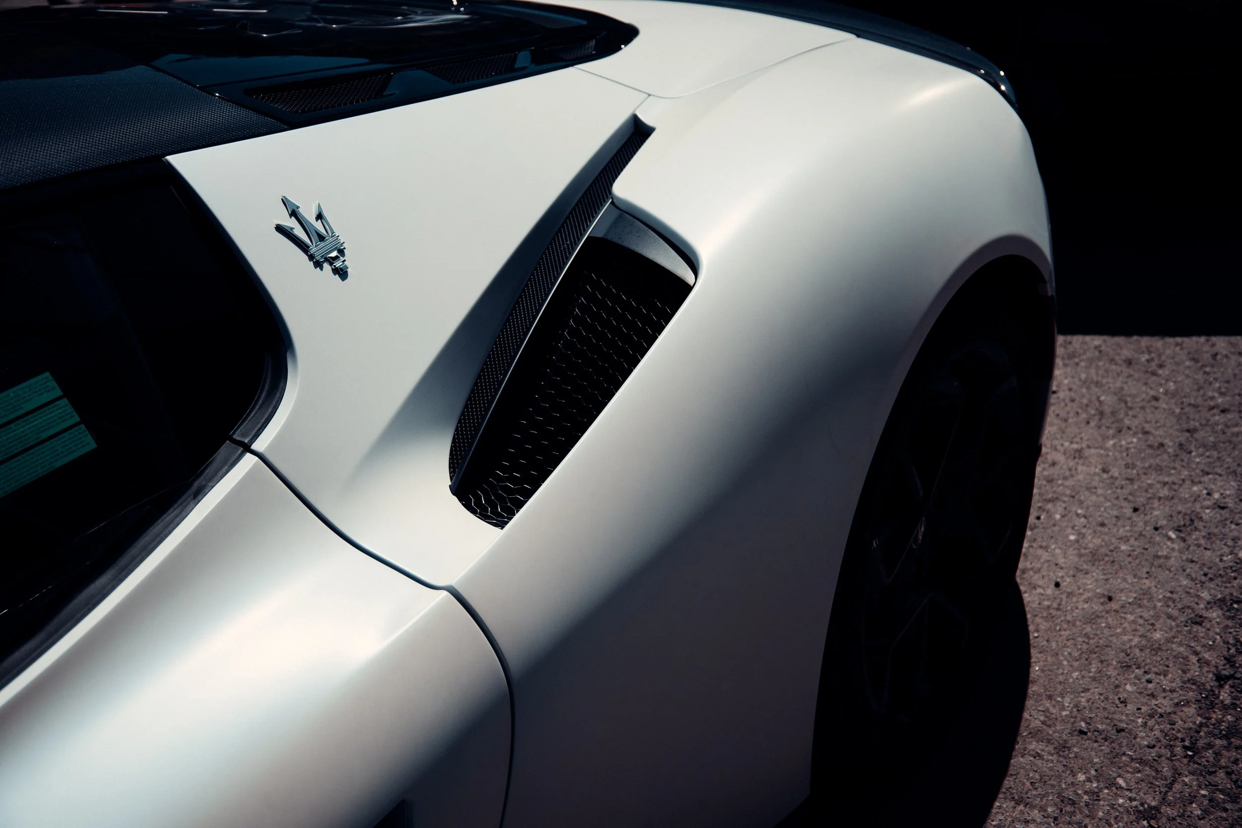 Close-up of the front part of a white Maserati sports car, showing the Maserati emblem and air vent on the hood, with part of the front wheel visible on the right.