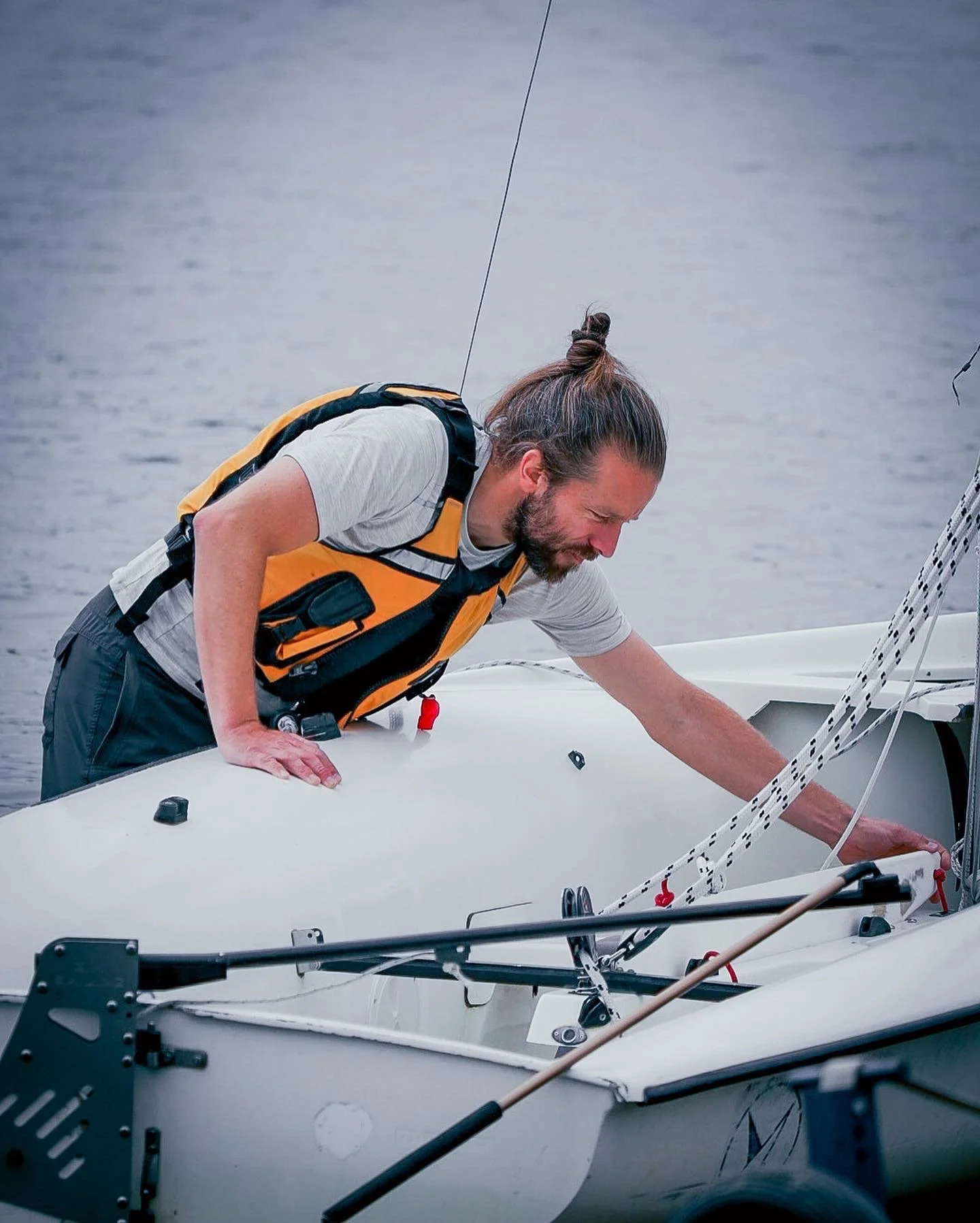 A man with a beard and long hair tied back, wearing a life jacket, leaning over the edge of a small sailboat on a body of water, adjusting the boat's rigging.