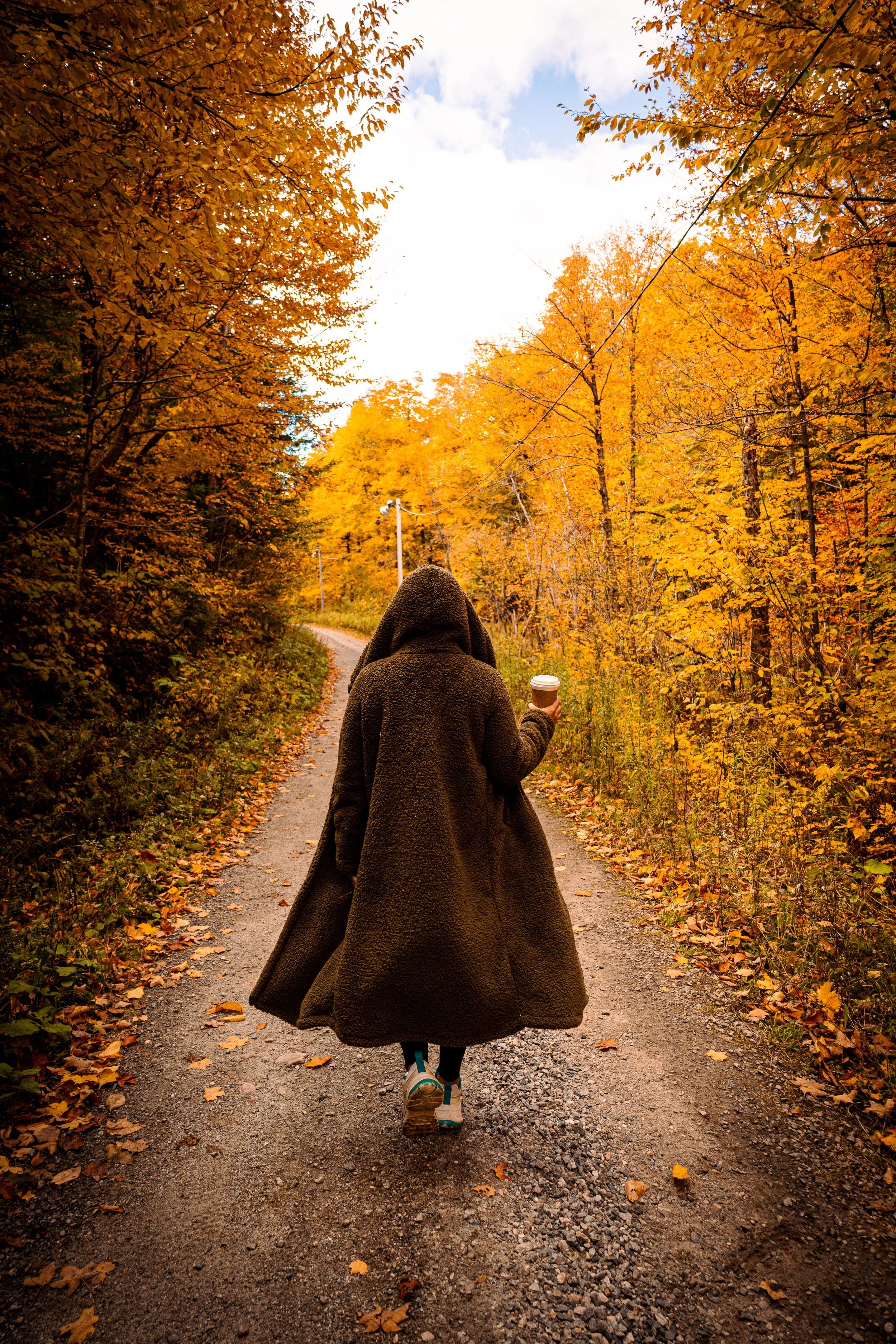 Person wearing a brown coat and sneakers walking on a dirt road through a forest with orange and yellow fall foliage, holding a coffee cup.