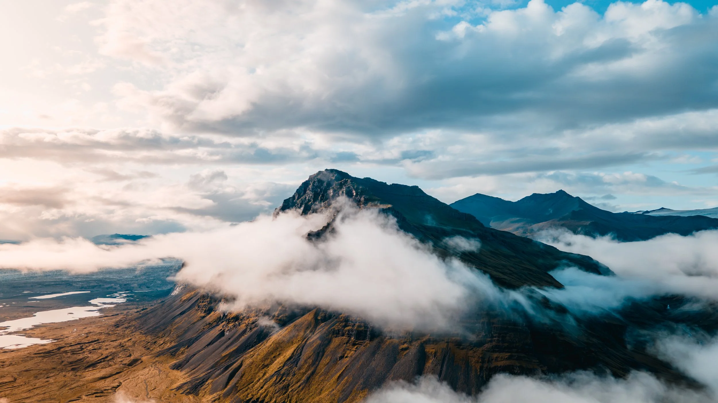A mountain landscape with clouds surrounding rugged peaks and green slopes under a partly cloudy sky.