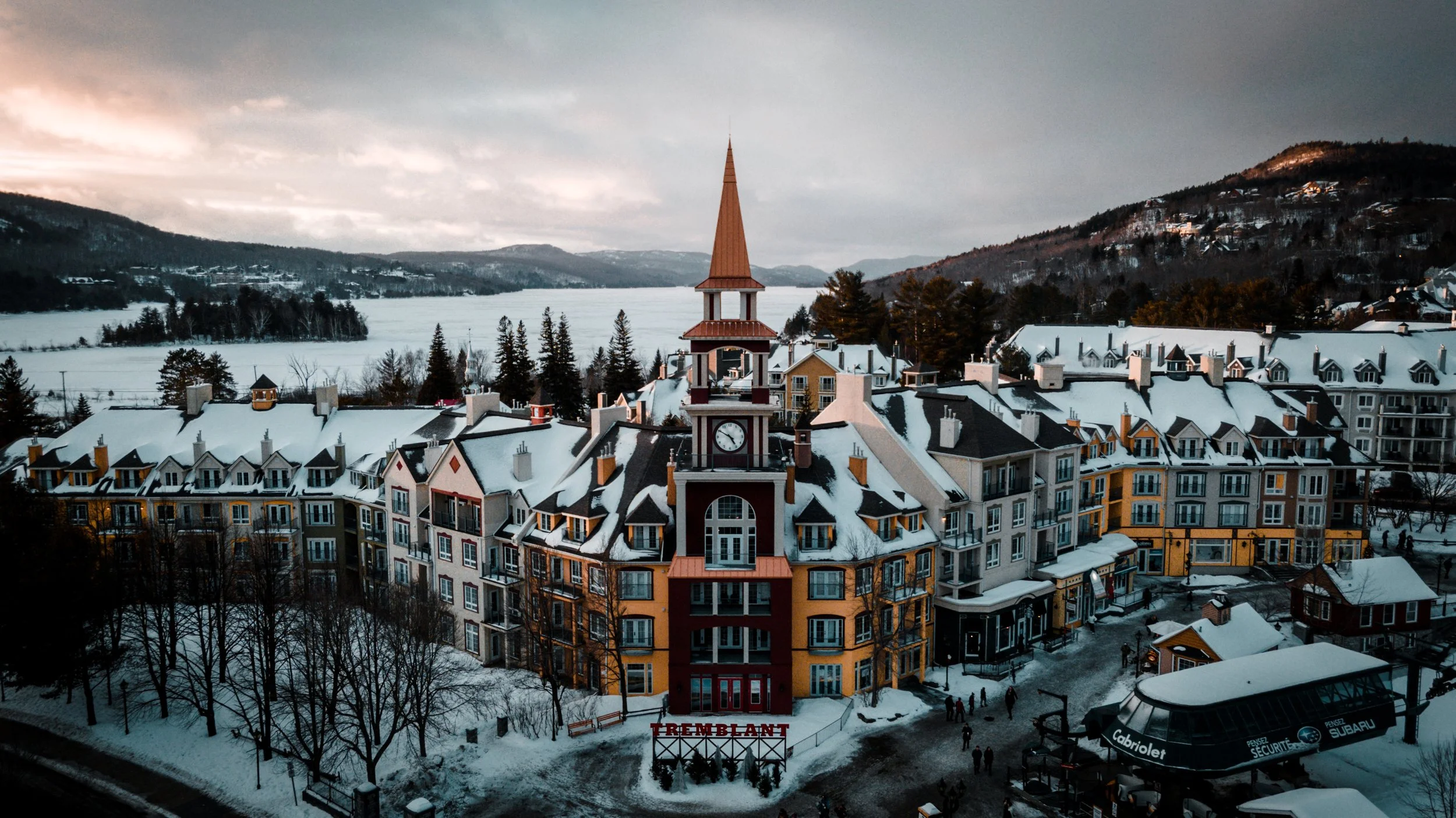 A snowy mountain town features a large, colorful building with a prominent clock tower and red doors, with the word 'TREMPLANT' displayed at the front. Surrounding the building are smaller structures, leafless trees, and a ski lift in the foreground.