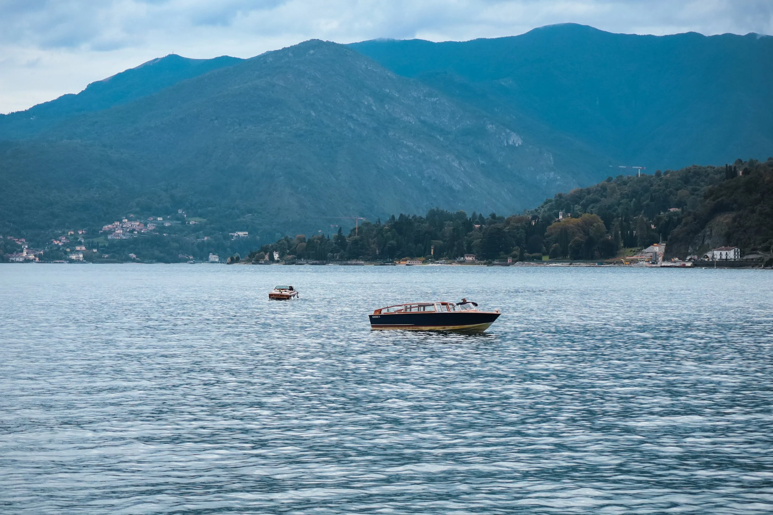 A scenic view of a lake with three boats floating on the water, surrounded by lush green mountains under a cloudy sky.