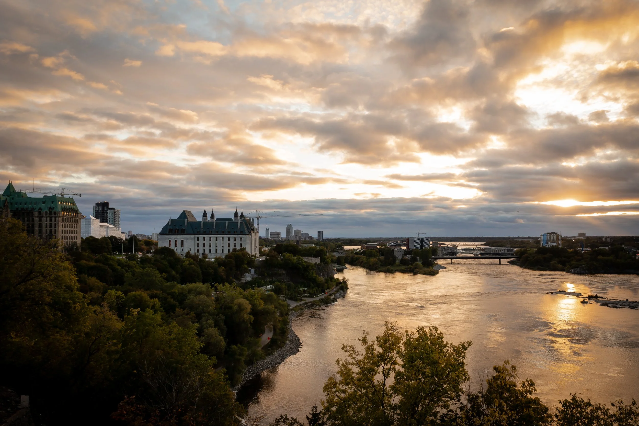 Sunset over a river with buildings and trees along the shoreline, cloudy sky with patches of sunlight.