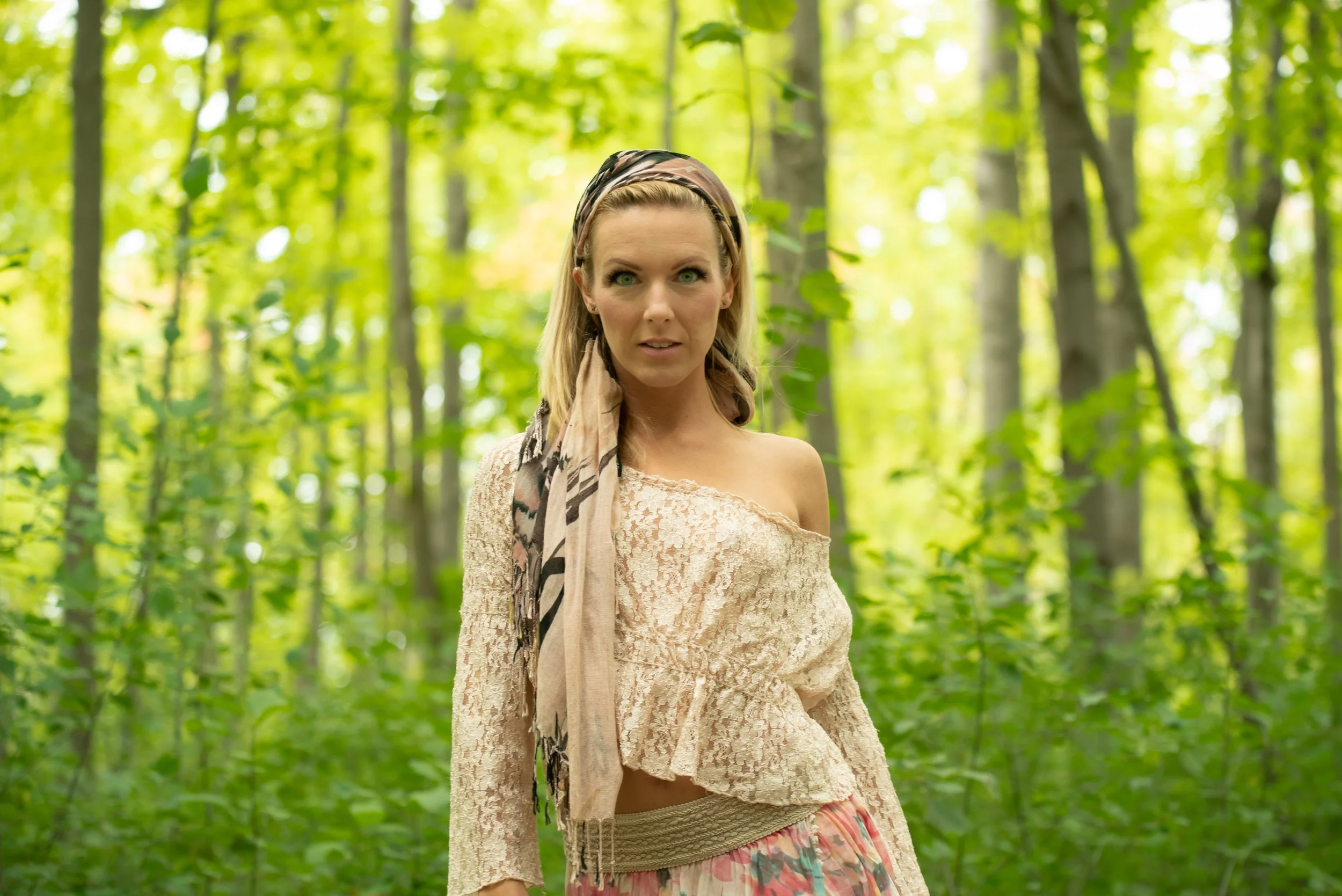 A woman standing in a lush green forest, wearing a beige off-shoulder lace top and a multicolored skirt, with a patterned headscarf.