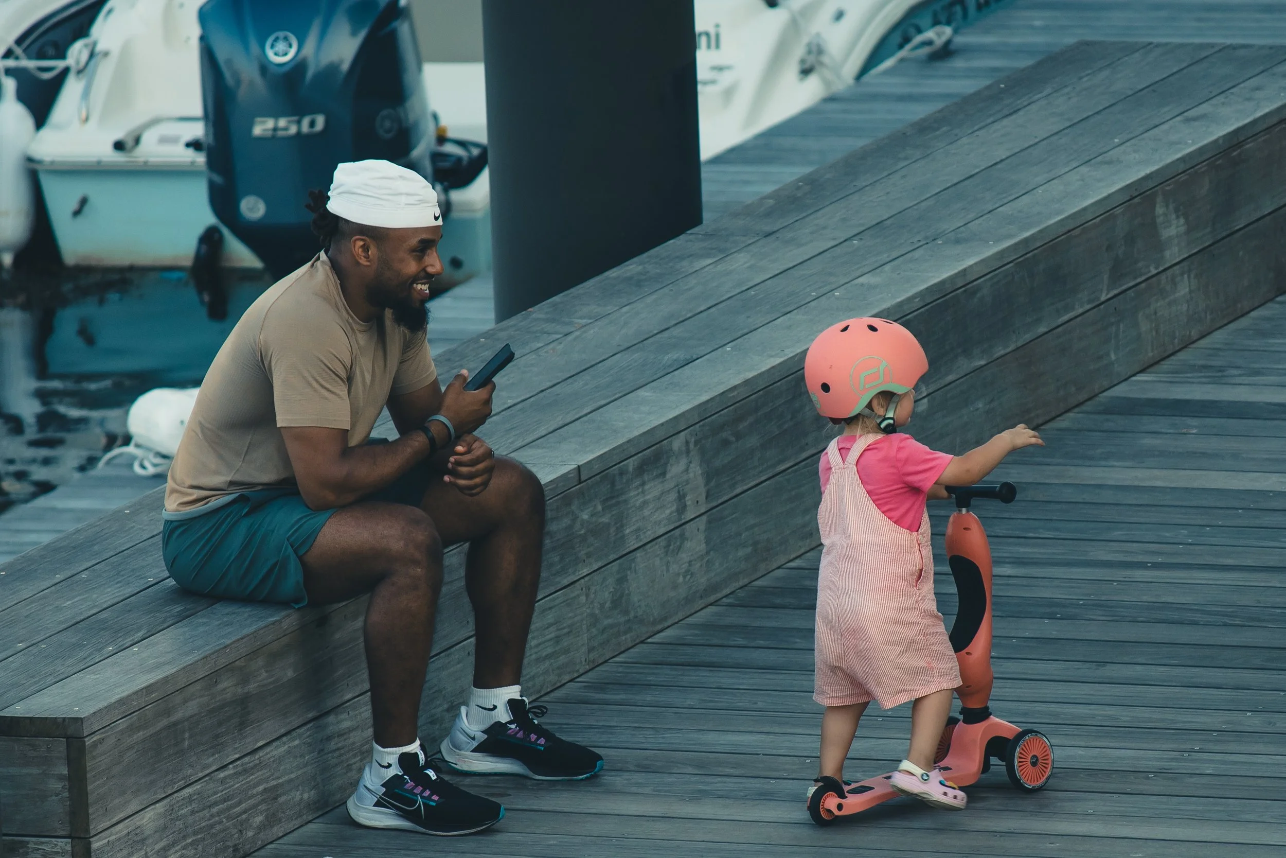 A man is sitting on a wooden dock, smiling and looking at a young girl on a scooter. The girl is wearing a pink helmet and pink outfit, and is riding a pink scooter near boats docked at the marina.