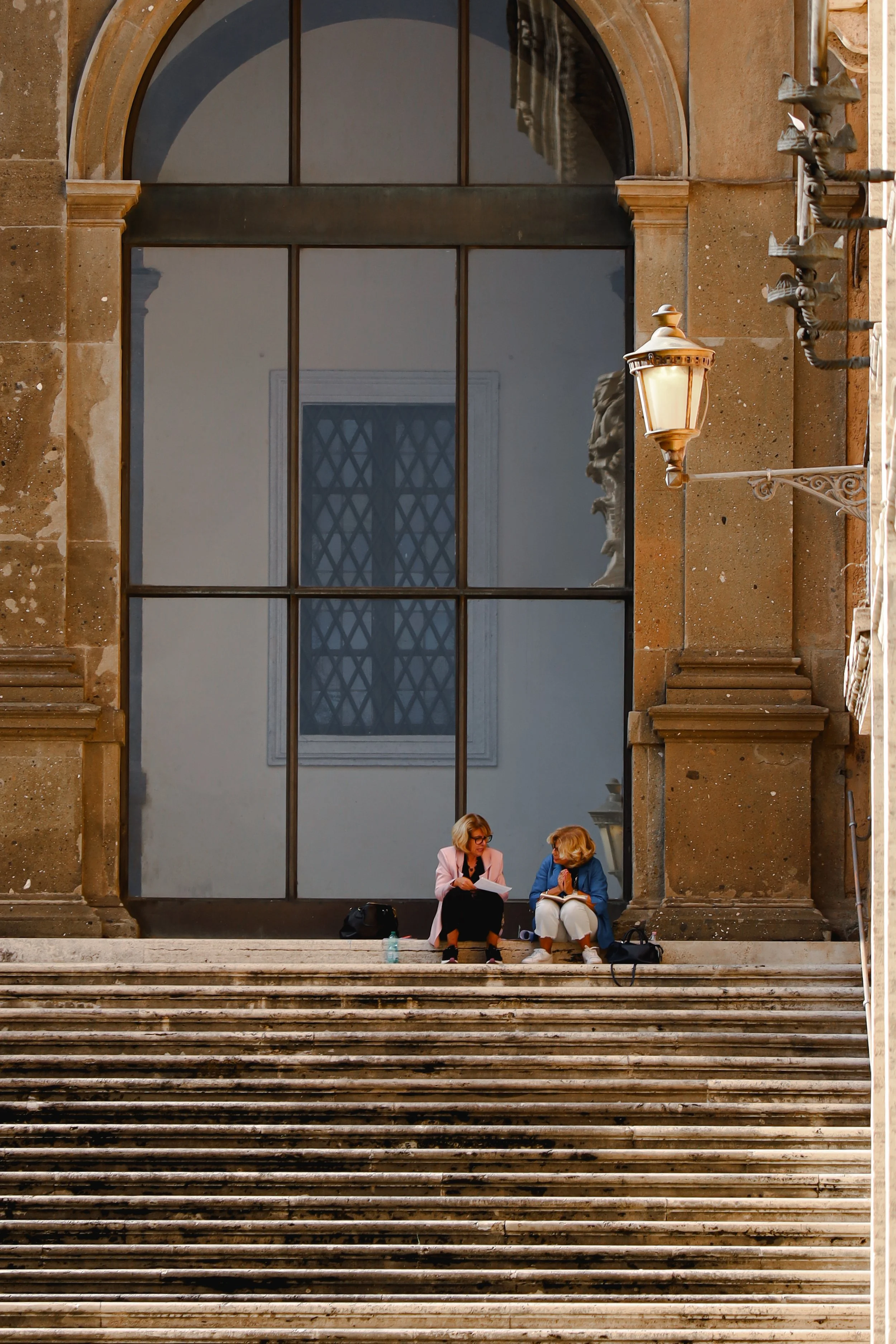 Two women sitting on steps in front of a large window, engaged in conversation, with bags and water bottles nearby, in front of an historic building with ornate stone architecture and a street lamp.