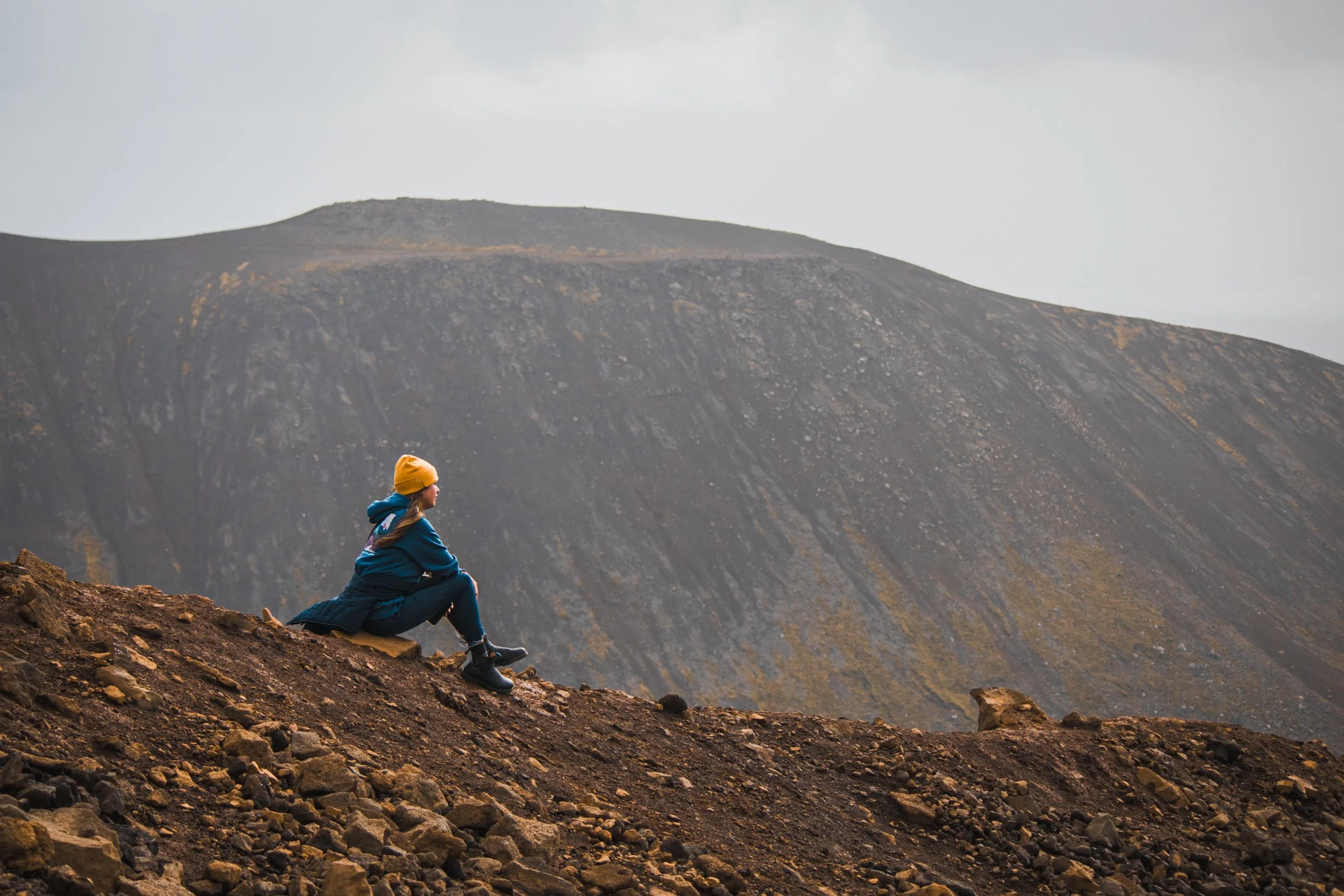 A person sitting on rocky terrain in front of a large, barren mountain, wearing a blue jacket, yellow beanie, and black boots.