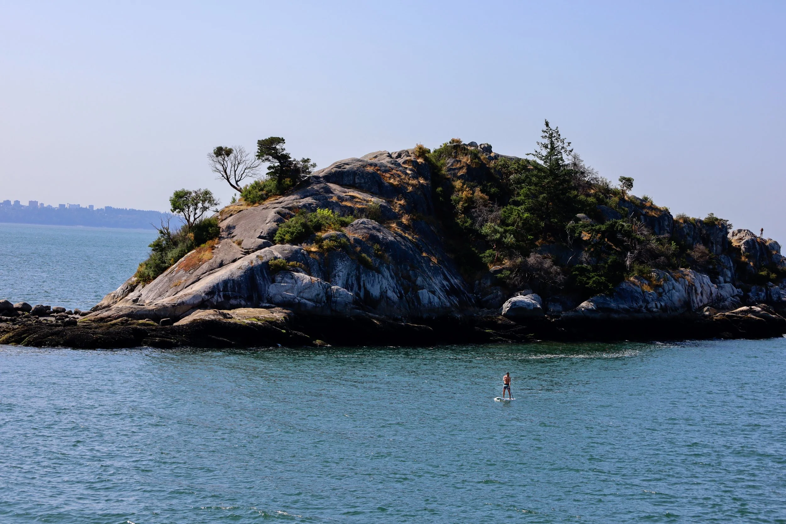 An island with rocky terrain and sparse trees, surrounded by water, with a person paddleboarding near the shore.