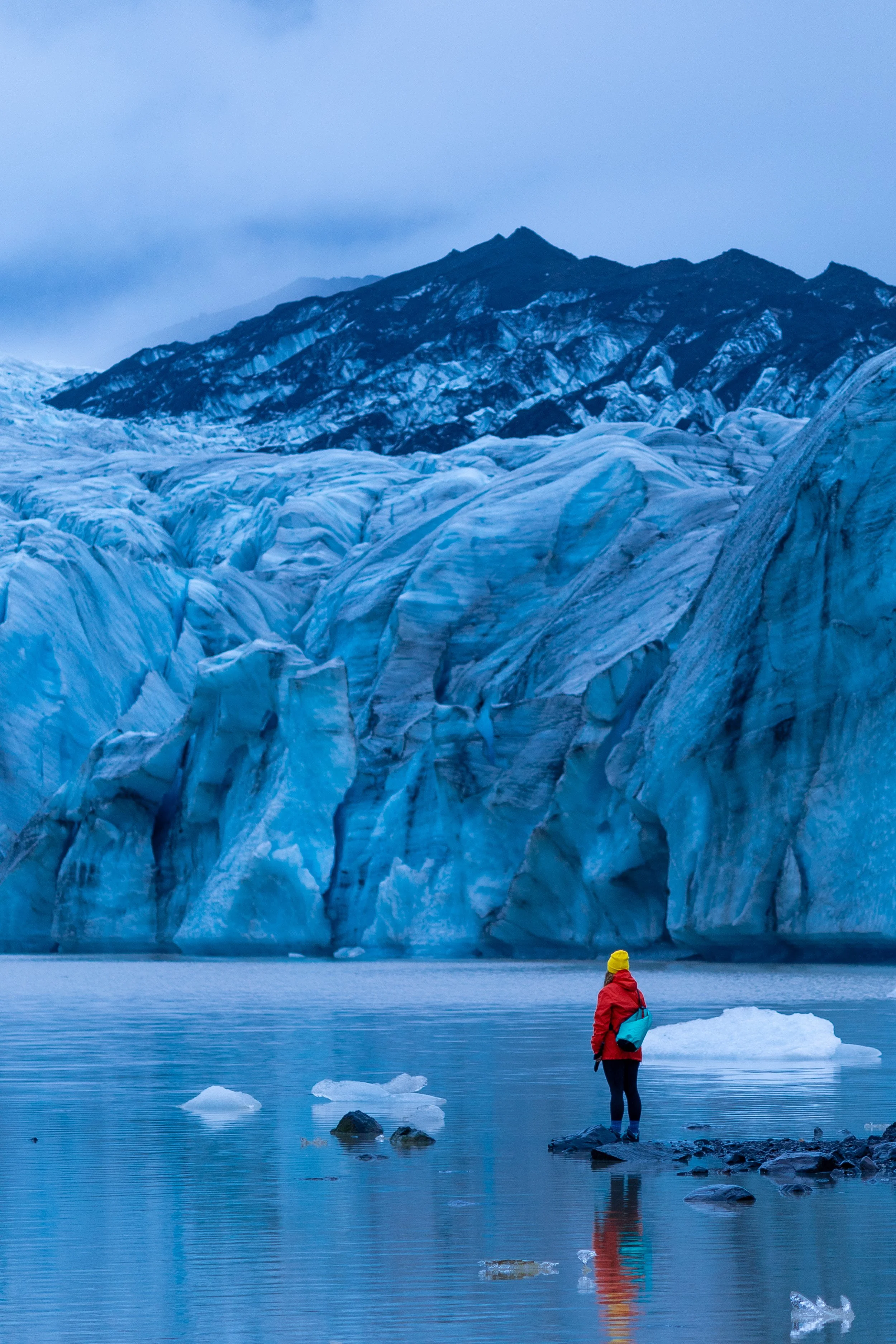A person in a red jacket and yellow hat standing near icy water with glaciers and mountains in the background.