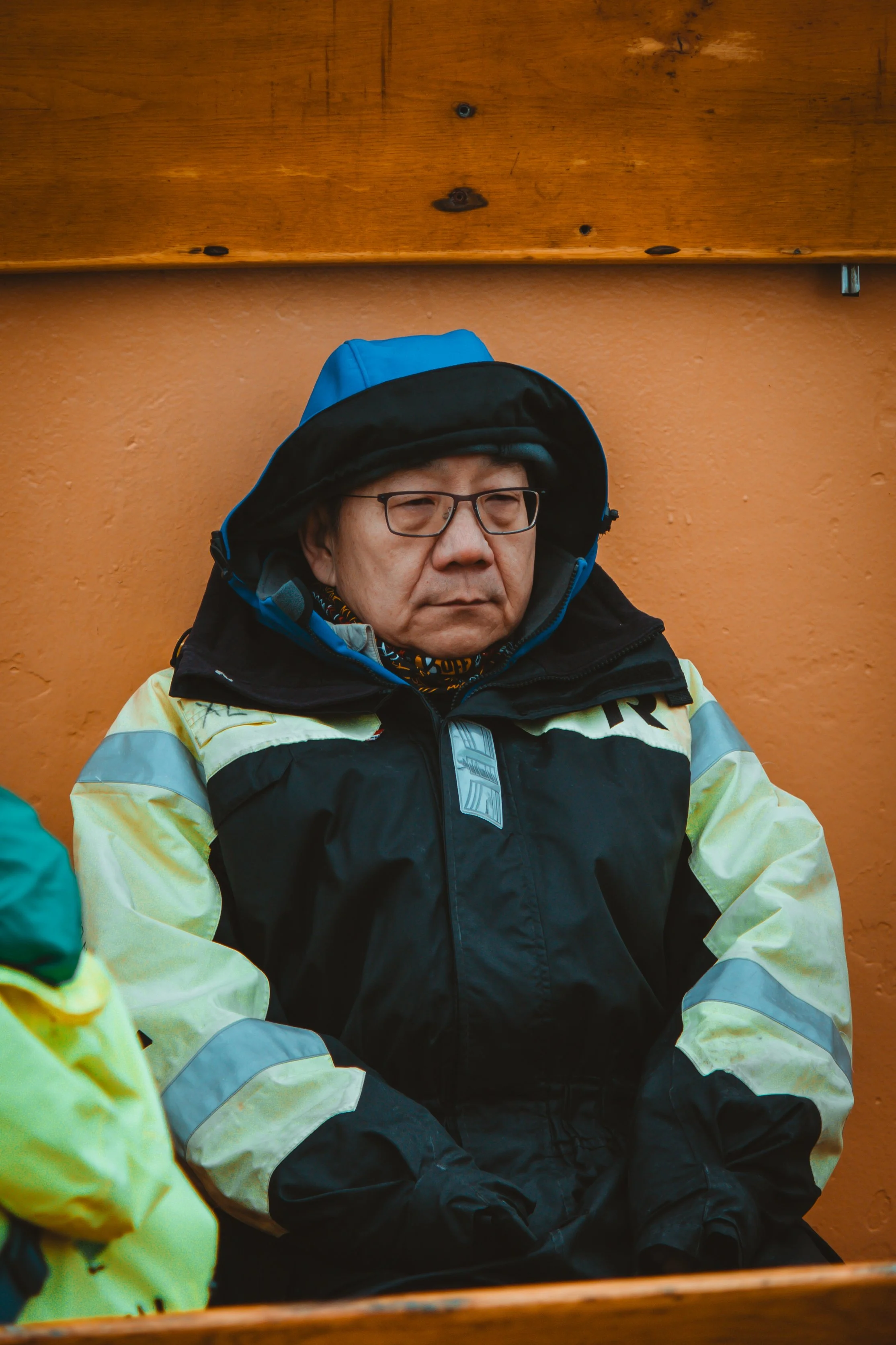 Man wearing black and yellow high-visibility jacket and glasses, sitting against an orange wall, with a hood on.