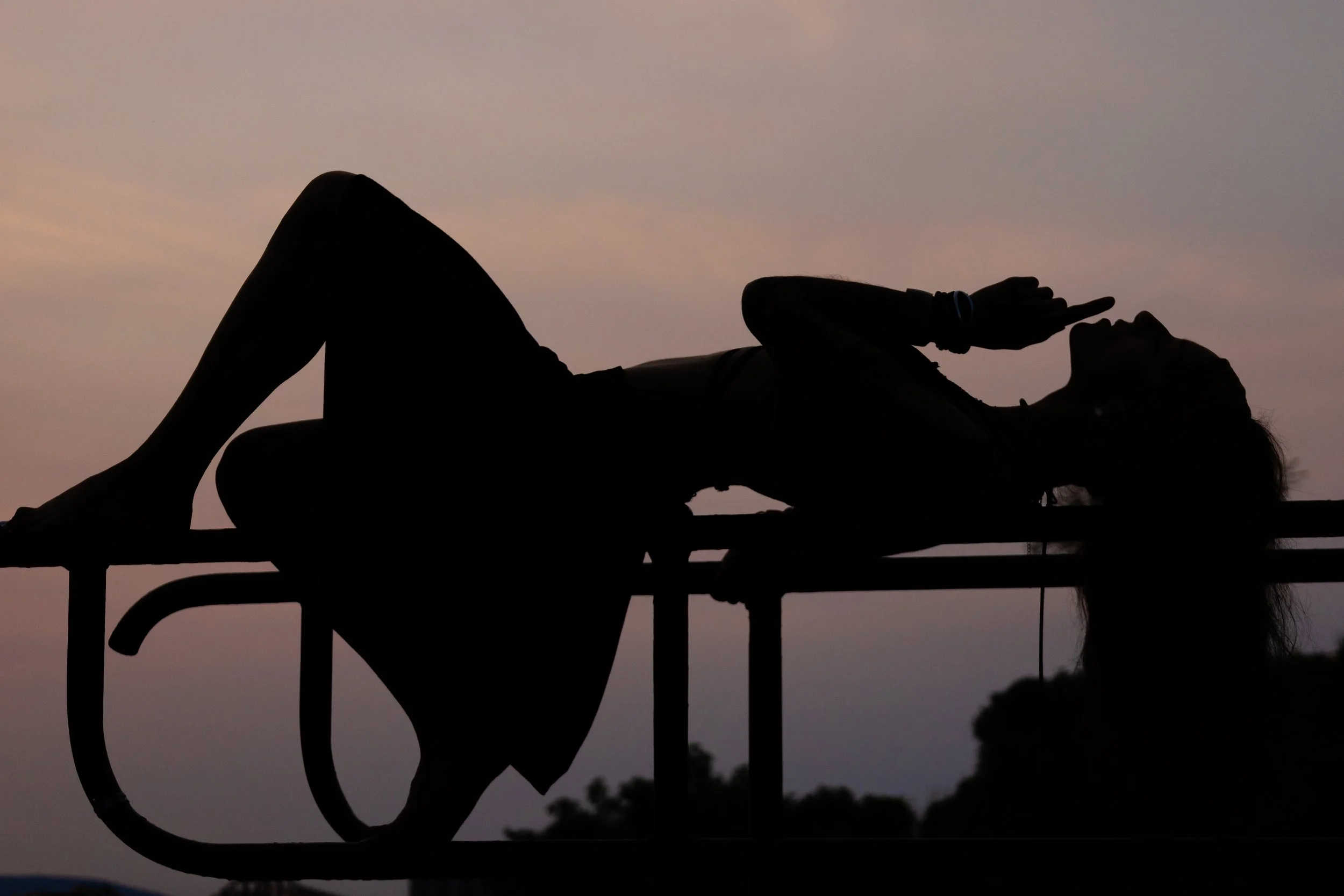 Silhouette of a woman lying on her back on a platform during sunset, with her hand near her face and her eyes closed.