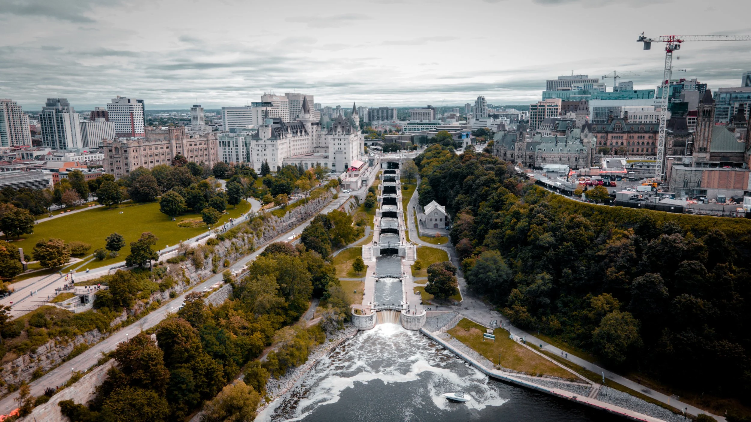 Aerial view of a Ottawa with a canal, waterfalls, green parks, and tall buildings, including construction cranes, under a cloudy sky.