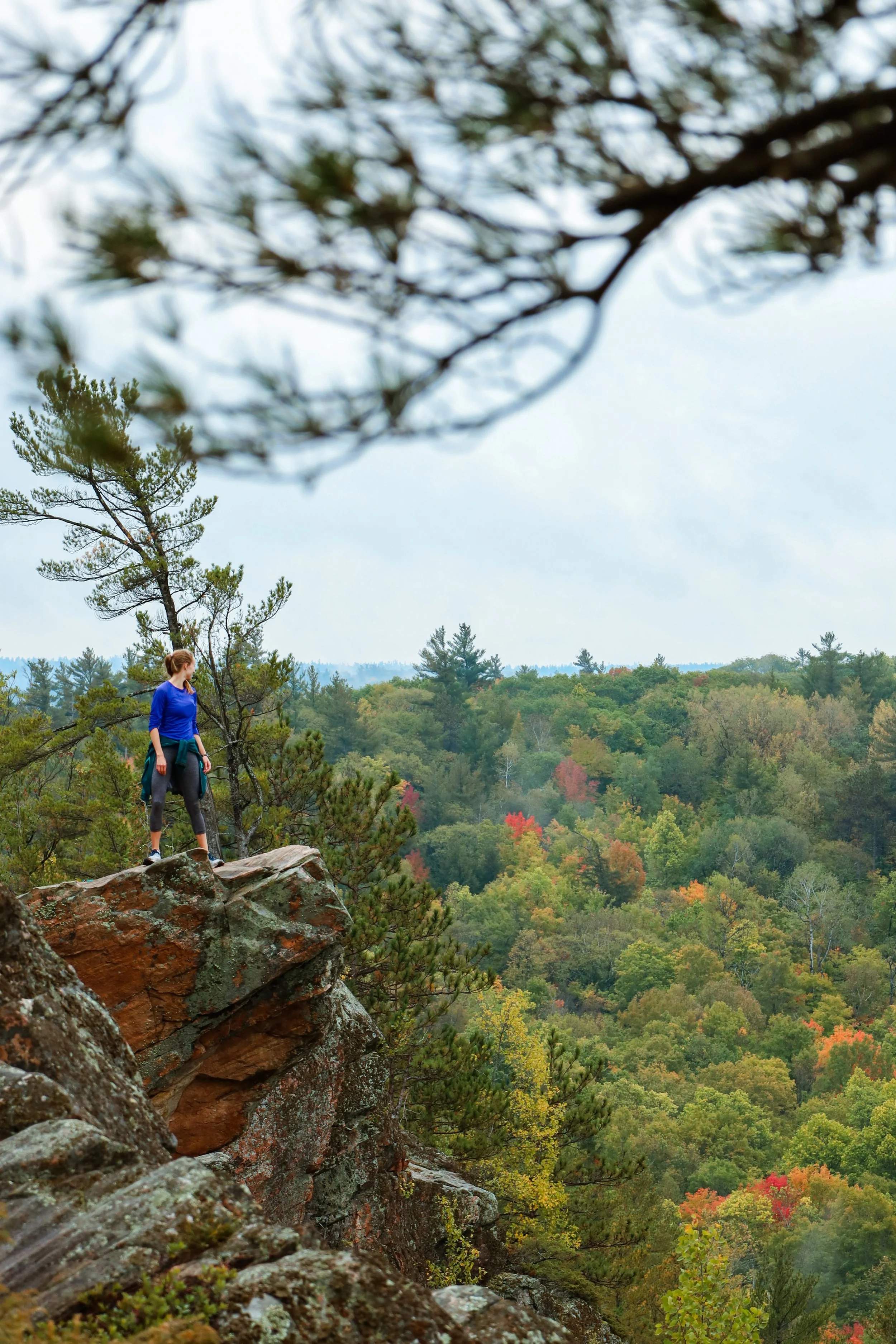 A woman standing on a rocky outcrop in a forested mountain area during autumn.