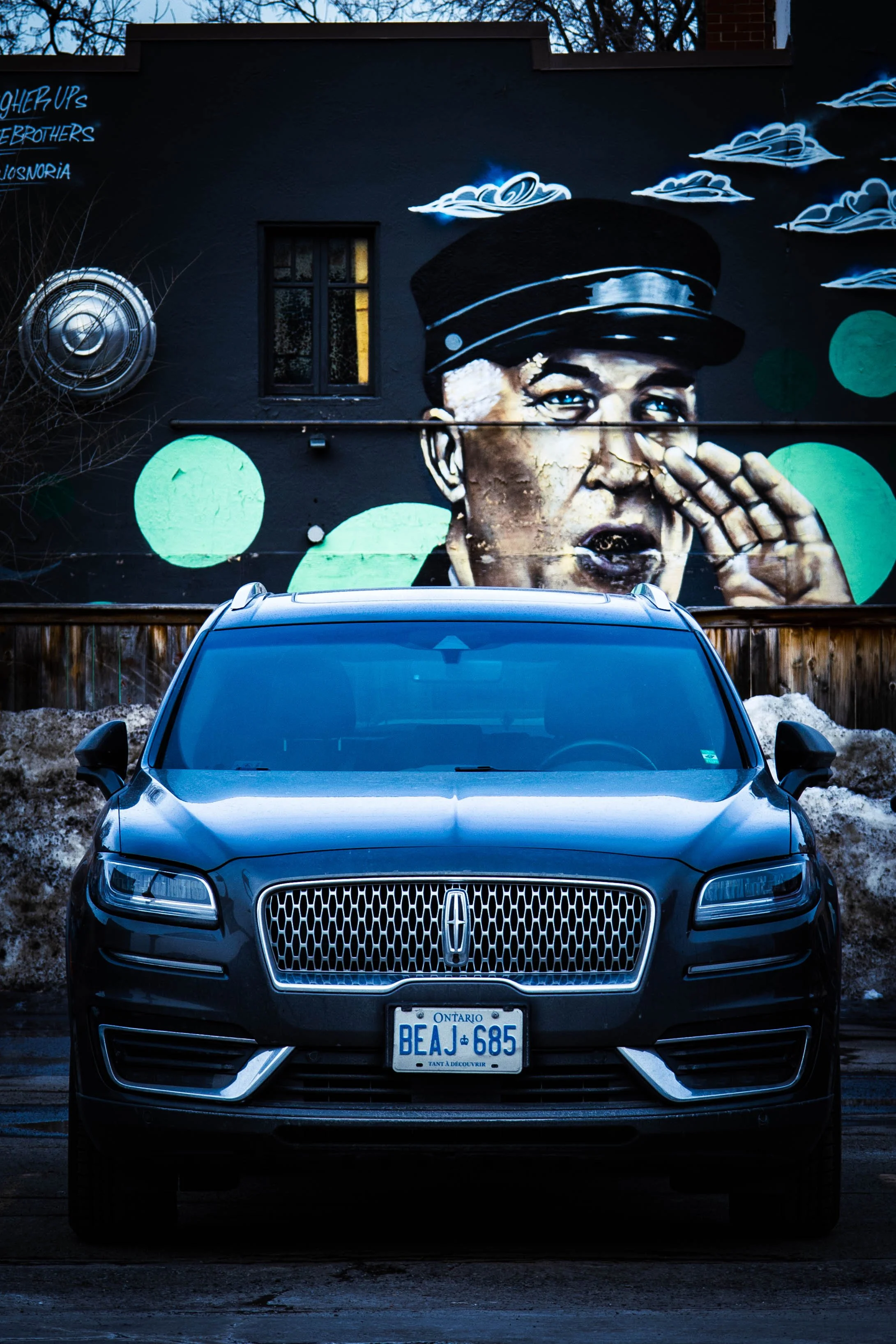 A black Lincoln SUV parked in front of a colorful mural of a person in uniform shouting into a megaphone, with clouds and abstract green circles in the background.