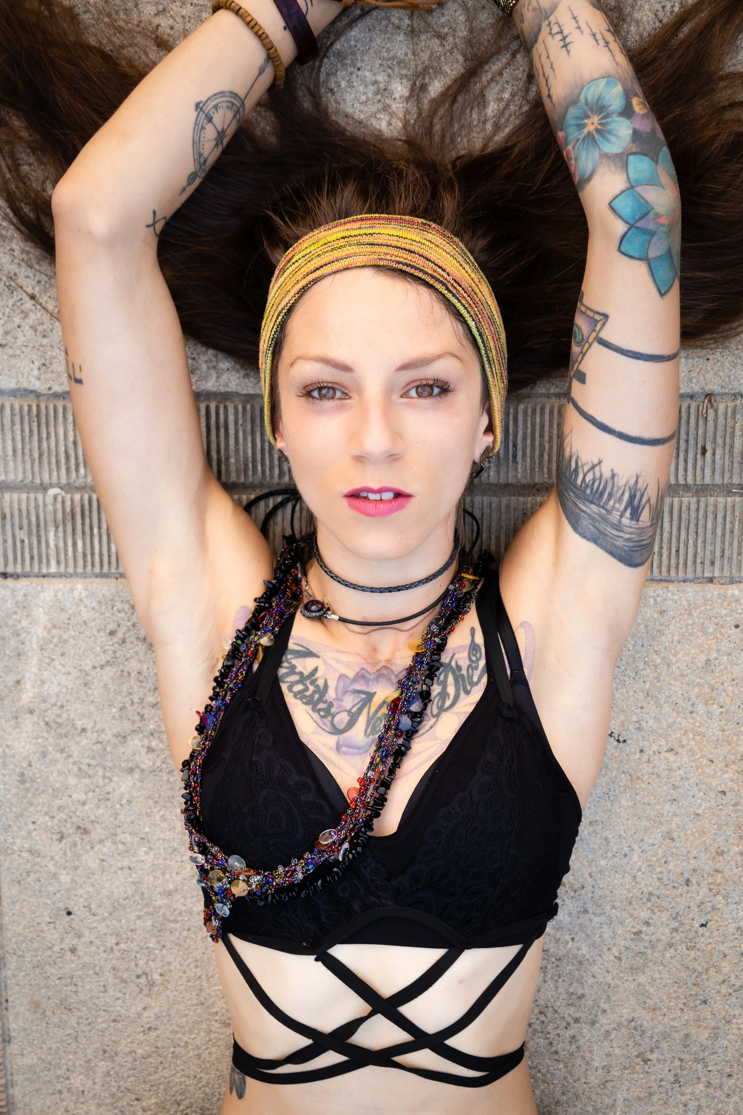 A young woman with tattoos, wearing a headband and layered necklaces, lying on her back on a concrete surface with her arms raised above her head.