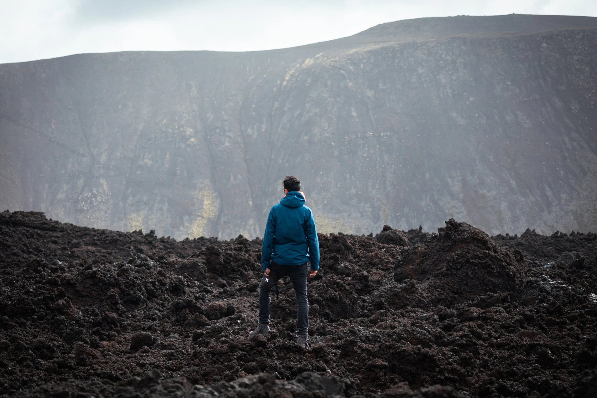 A person in a blue jacket standing on dark volcanic rocks, facing a large volcanic crater in the distance.